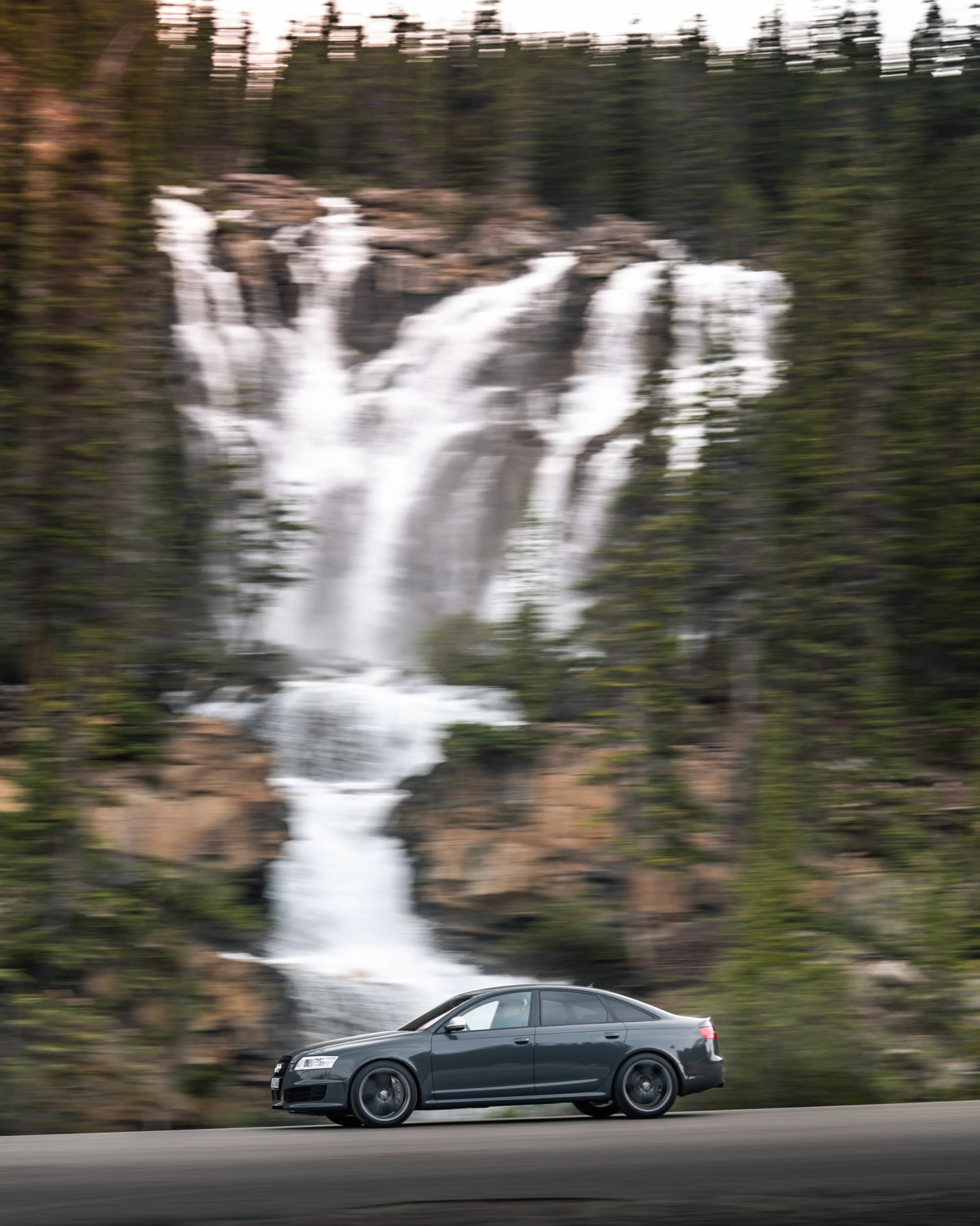 Dark gray sedan driving on a road with a large waterfall and forest in the background.