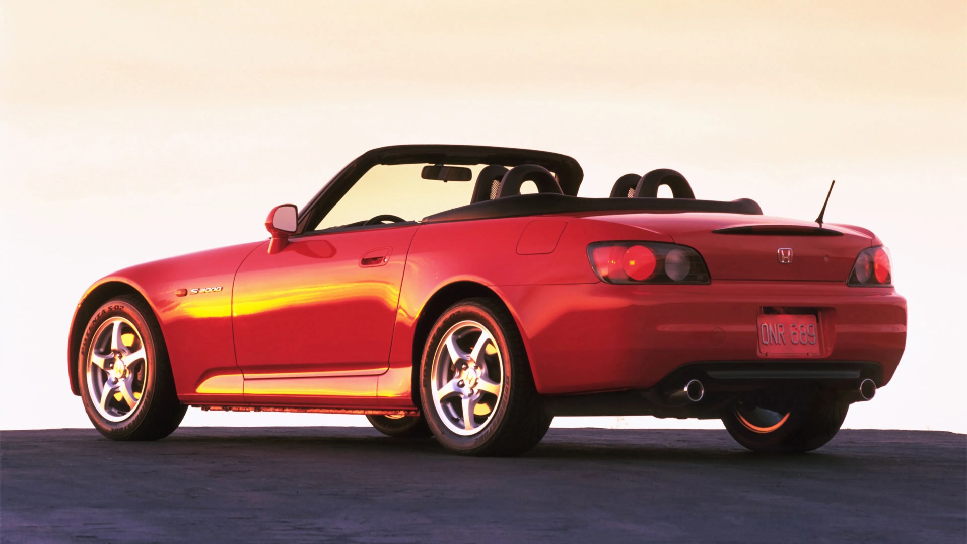Red Honda S2000 convertible sports car with black soft top down, shown from rear side on pavement.
