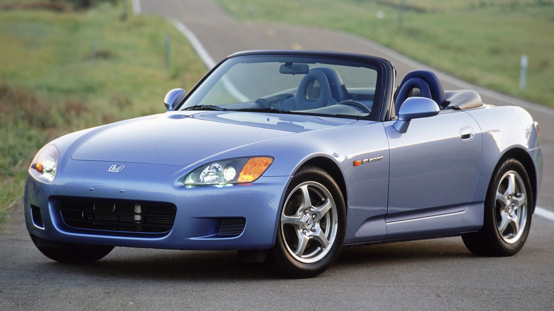 Light blue Honda S2000 convertible sports car parked on a road with a grassy background.