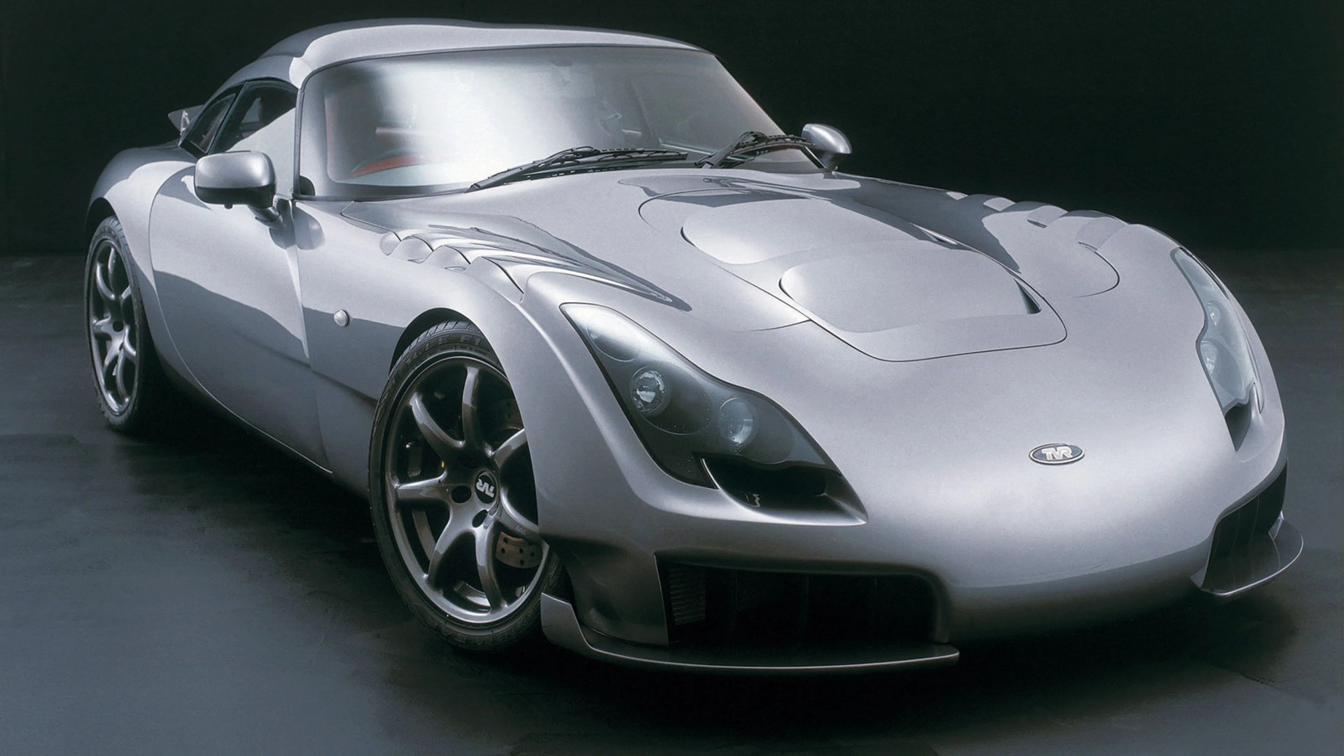 Silver TVR sports car with sleek aerodynamic design and dark alloy wheels on a dark floor.