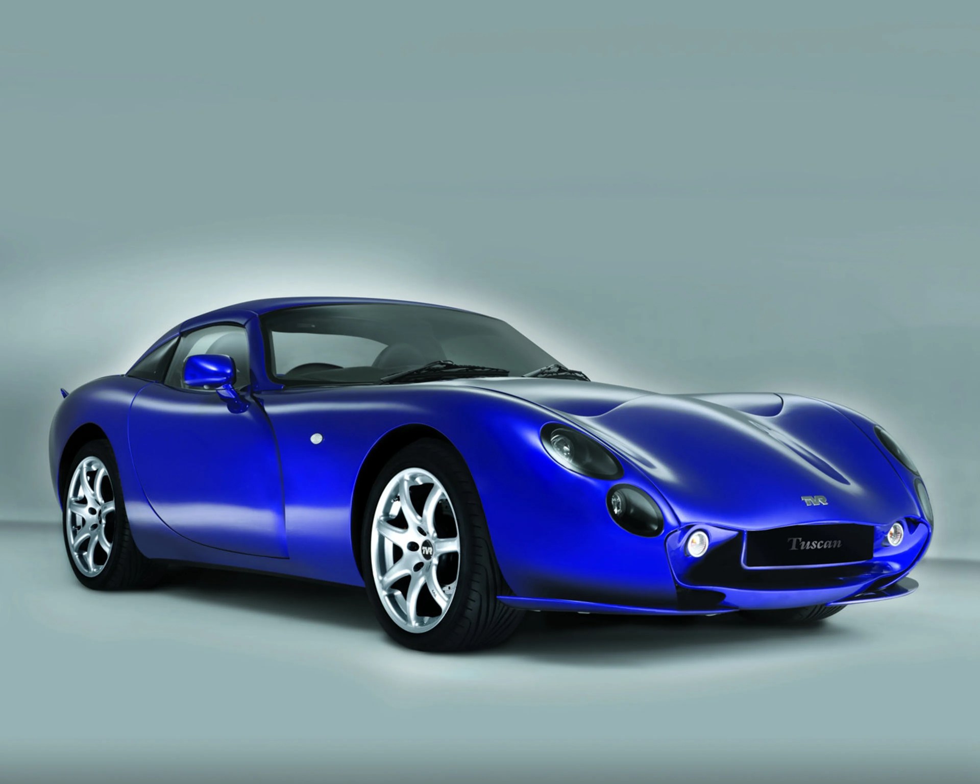 Blue TVR Tuscan sports car with sleek curves and silver alloy wheels on a gray background.