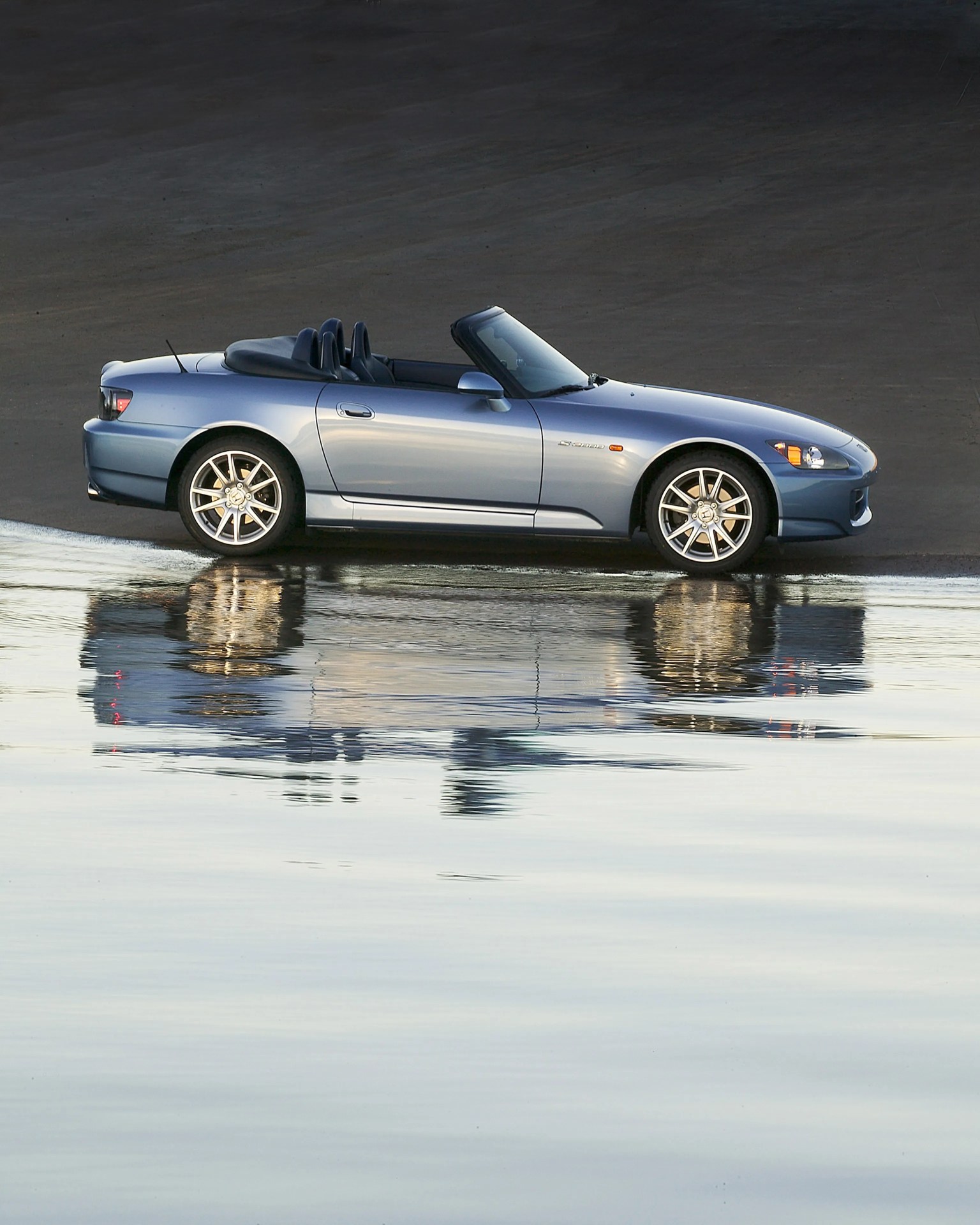 Blue convertible sports car parked on wet sand with its reflection in the water.