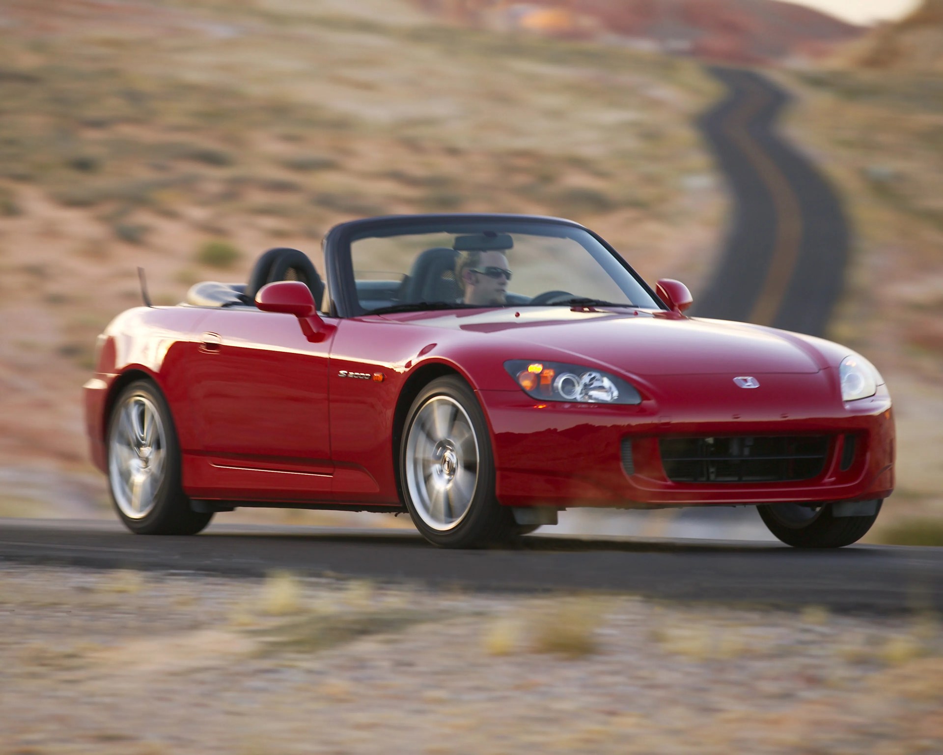 Red Honda S2000 convertible sports car driving on a winding desert road.