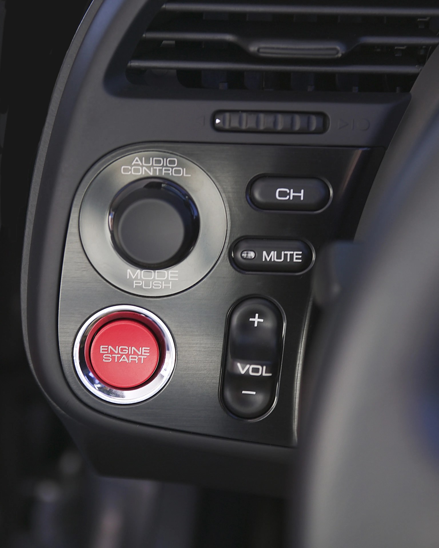 Close-up of a car dashboard panel with a red engine start button, audio control knob, channel and mute buttons, and volume controls.