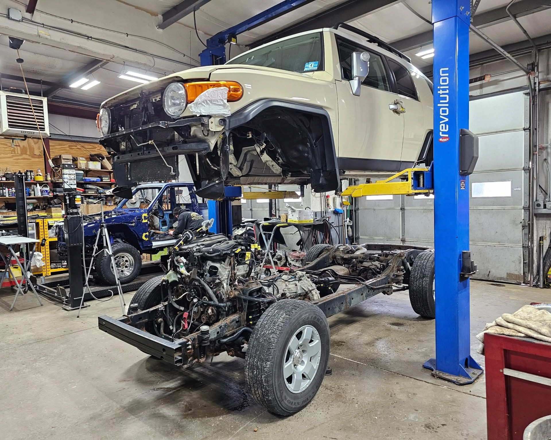 Beige SUV body lifted on a blue and yellow car lift with its chassis and engine below in a garage workshop.