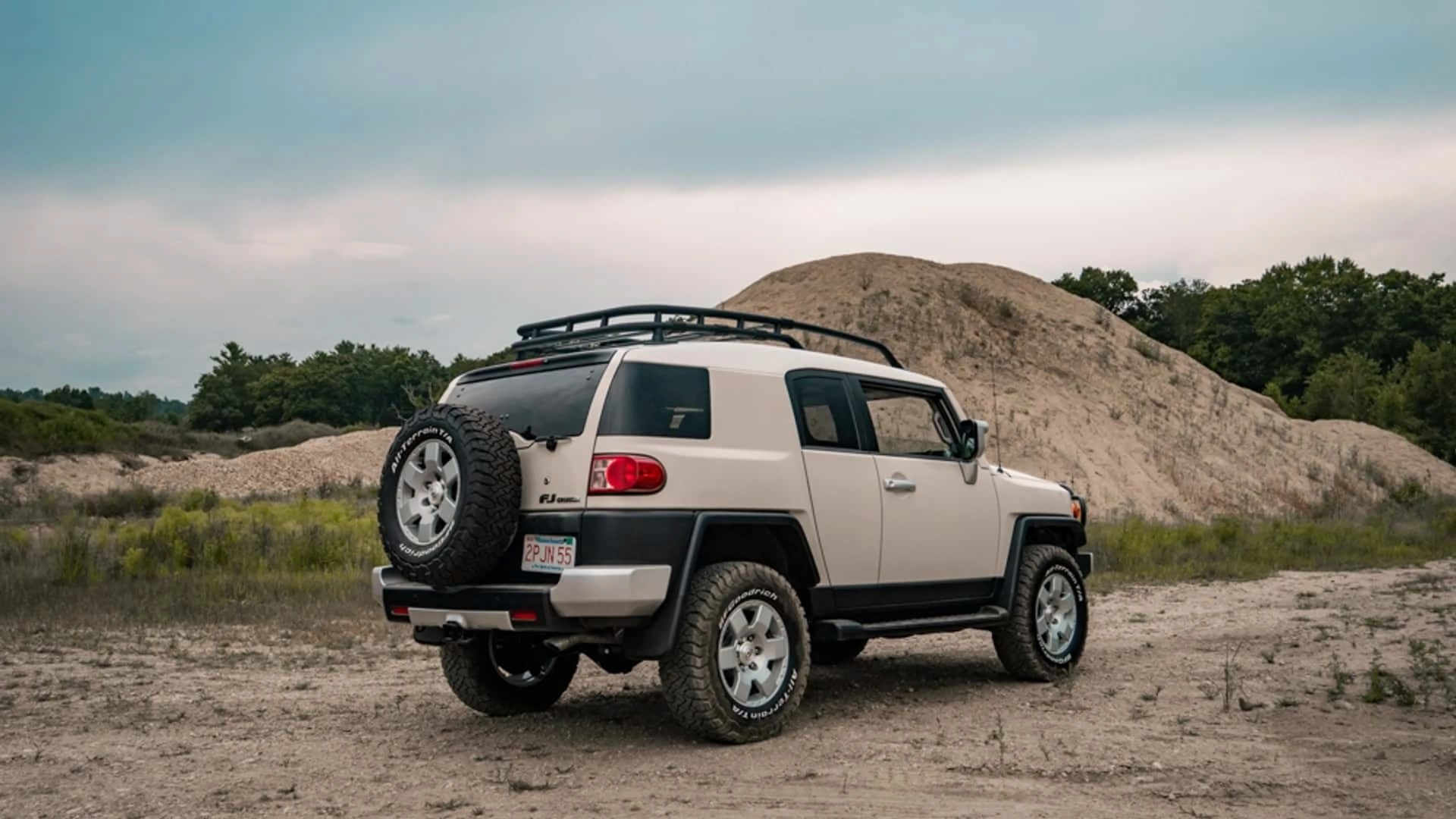 Beige Toyota FJ Cruiser with roof rack and spare tire on rear, parked on dirt with hill and trees in background.