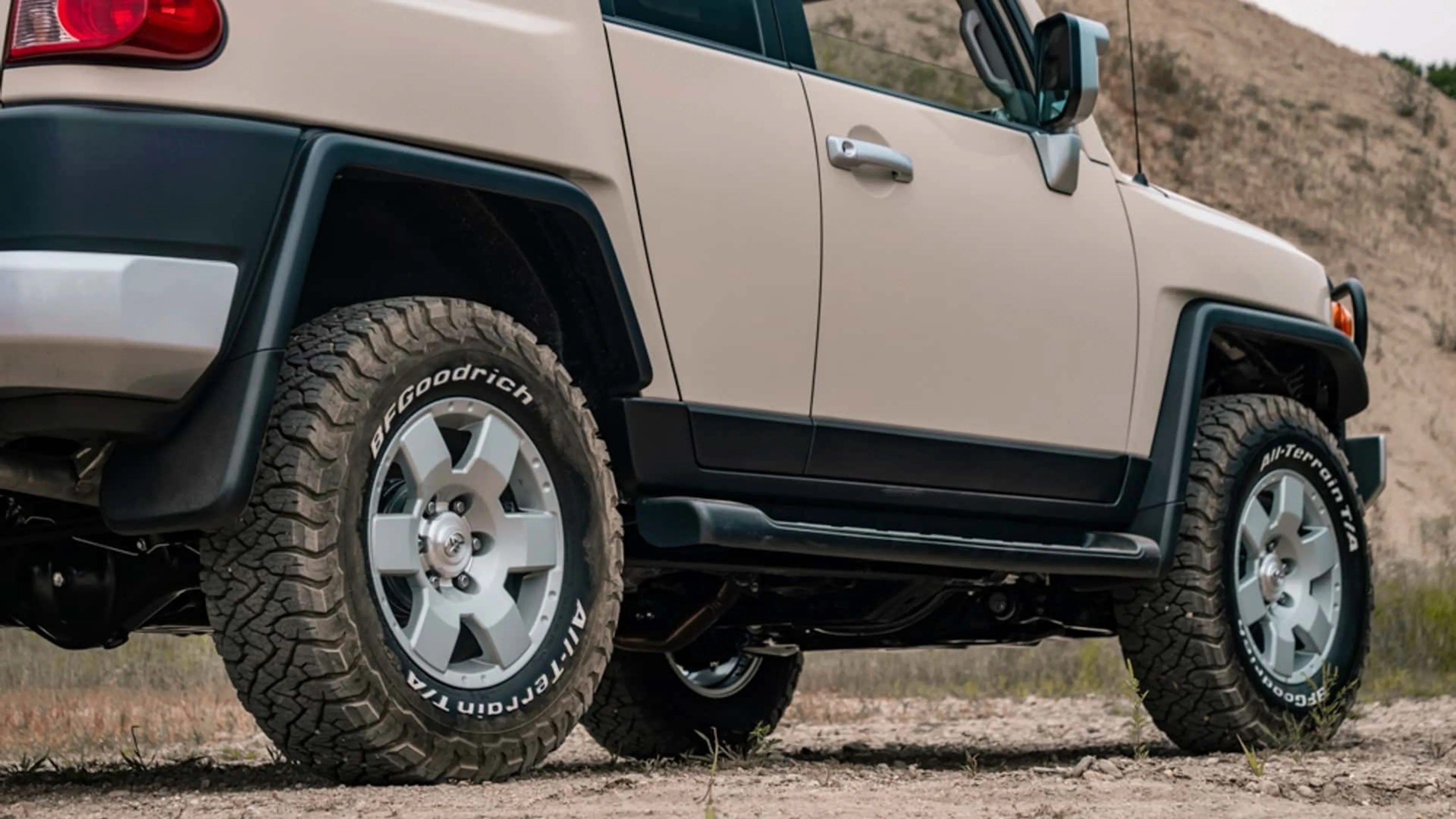 Close-up of beige off-road vehicle's side showing BFGoodrich All-Terrain T/A tires and silver alloy wheels on dirt ground.