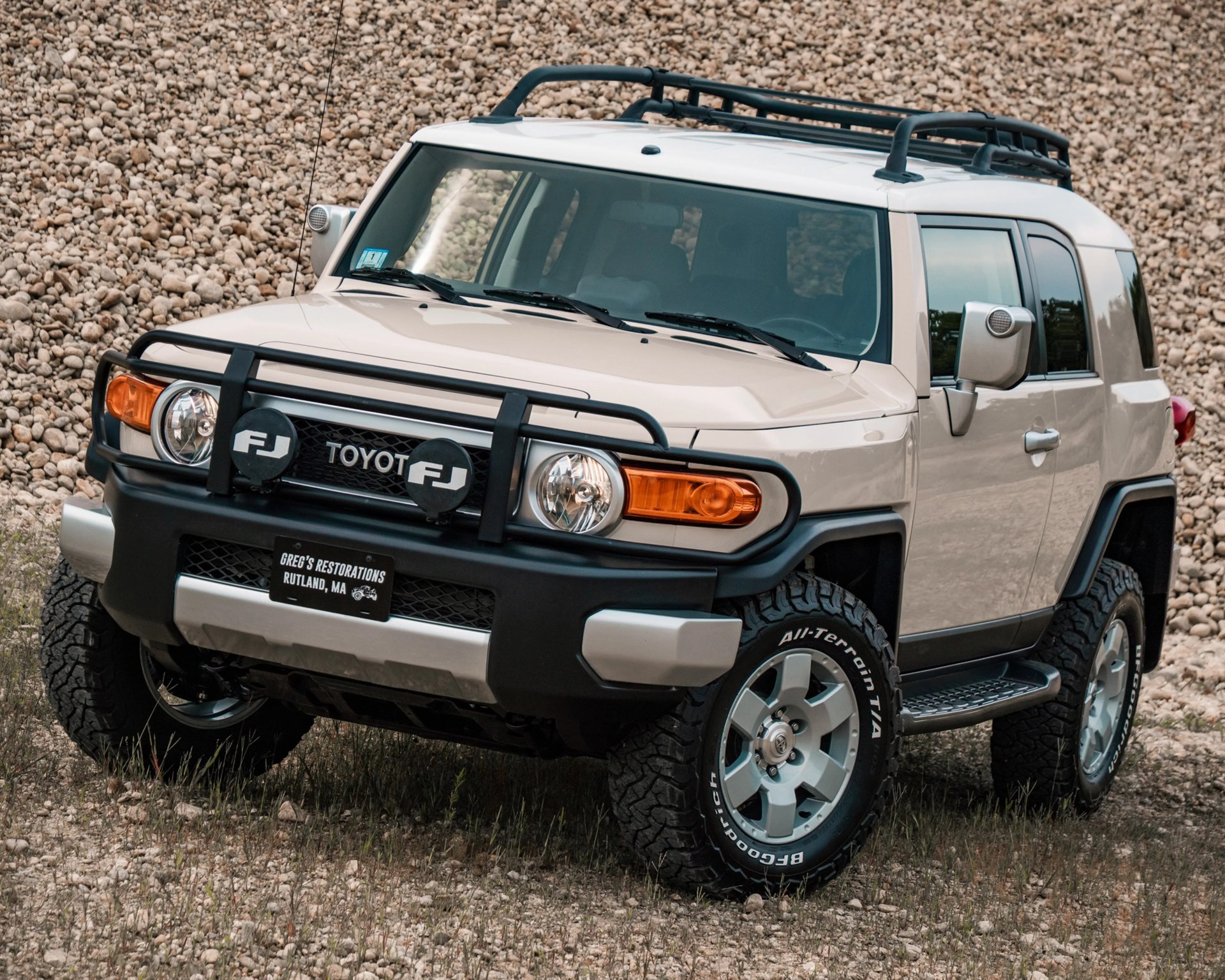 Beige Toyota FJ Cruiser with black roof rack and off-road tires parked on rocky terrain.