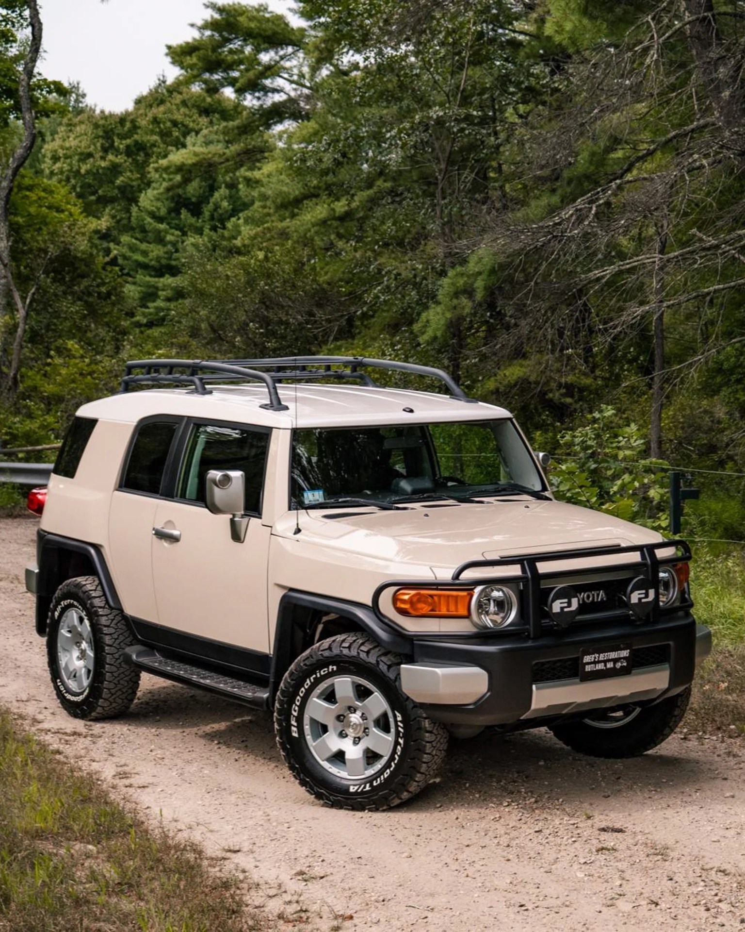 Beige Toyota FJ Cruiser with roof rack and off-road tires parked on a dirt path surrounded by trees.