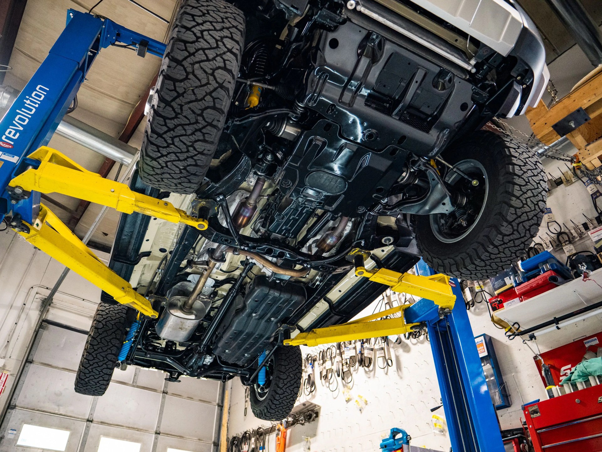 Underneath view of a lifted off-road vehicle showing suspension, exhaust, and tires on a blue and yellow hydraulic car lift in a garage.
