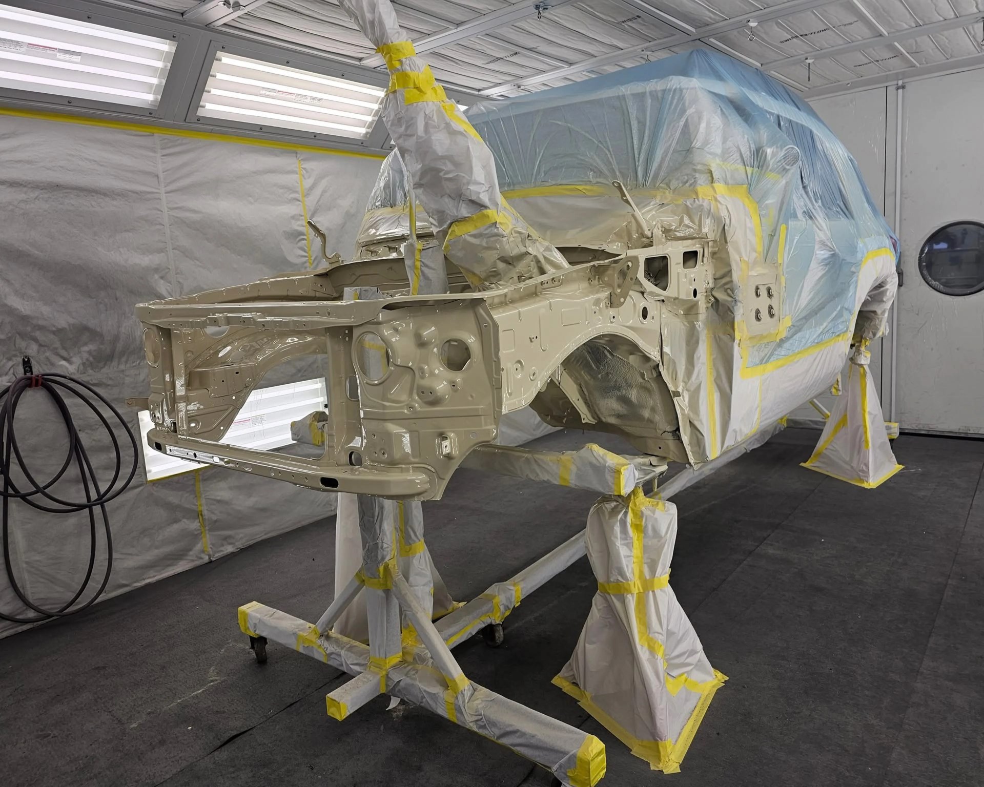 Car body frame painted beige and mounted on a stand covered with protective paper and tape in a paint booth.