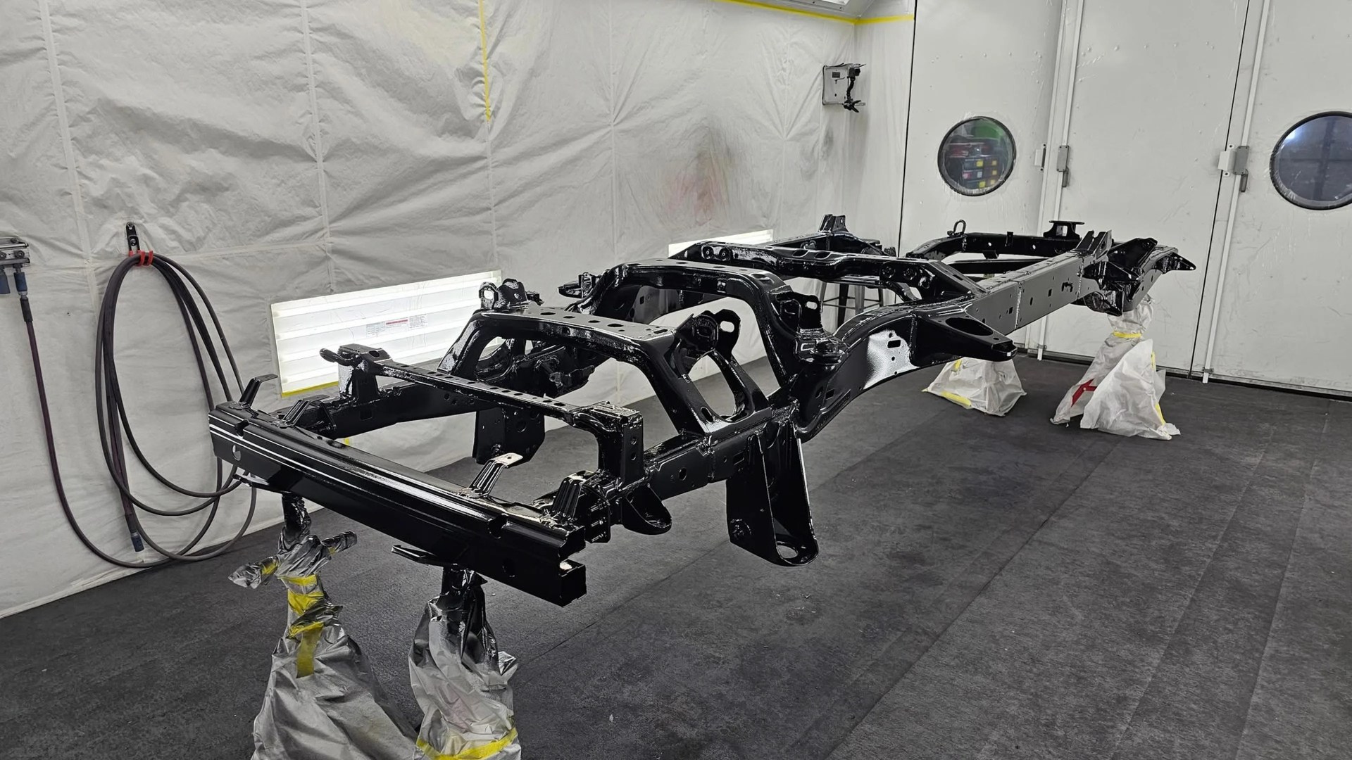 Glossy black painted vehicle chassis frame suspended on stands inside a white paint booth.