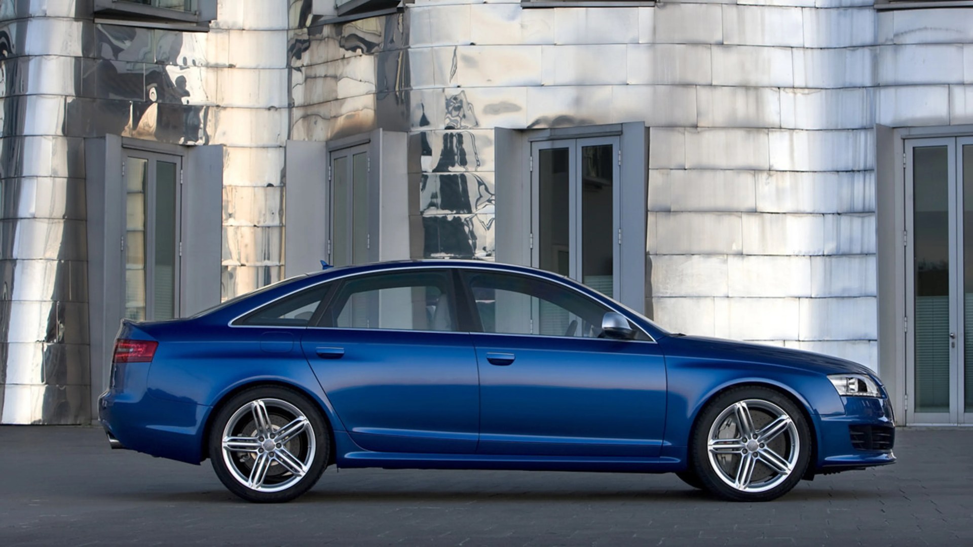 Blue luxury sedan with silver alloy wheels parked in front of a reflective metallic building.