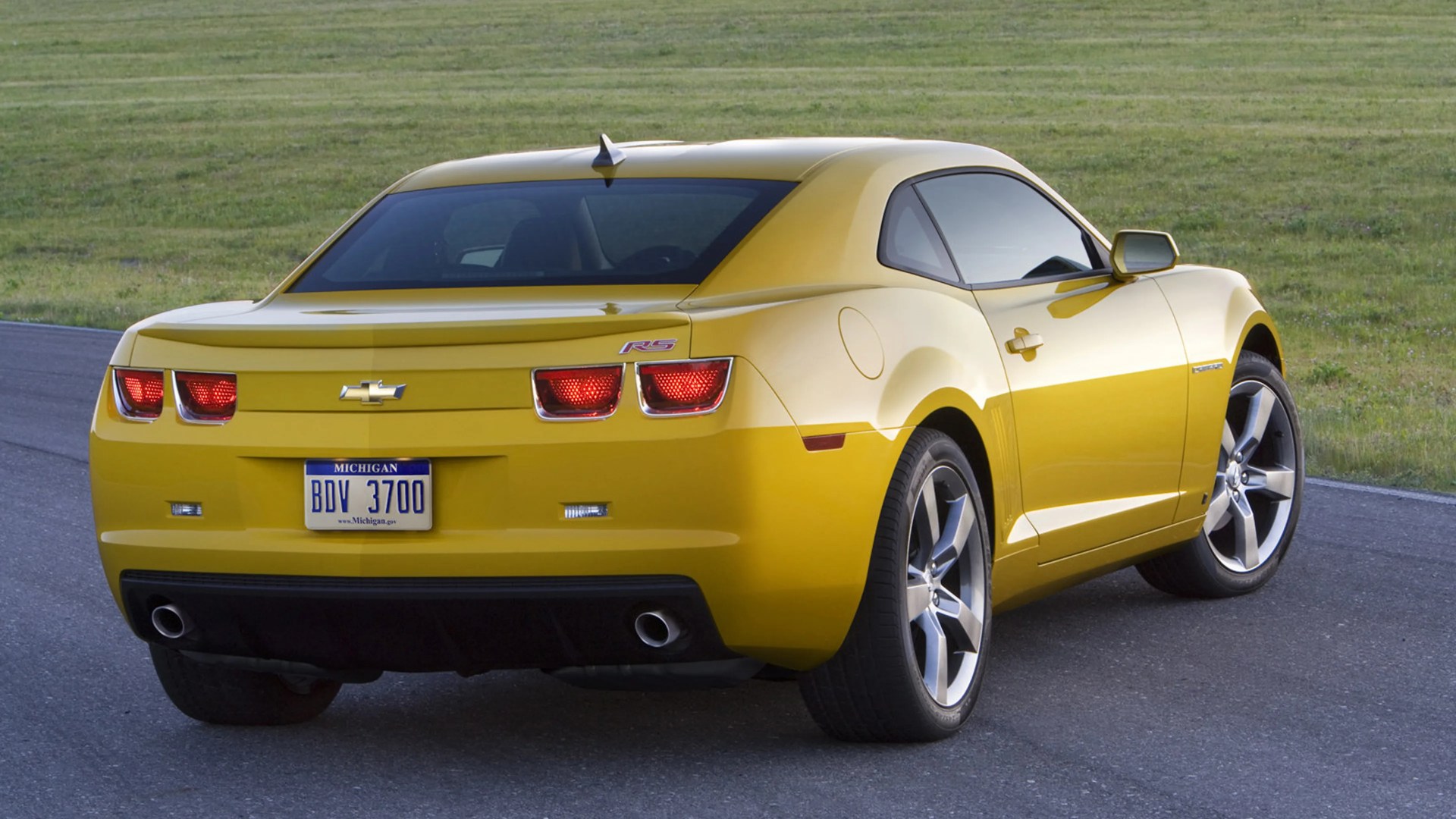 Yellow Chevrolet Camaro RS coupe with dual exhausts parked on a paved road near grass.
