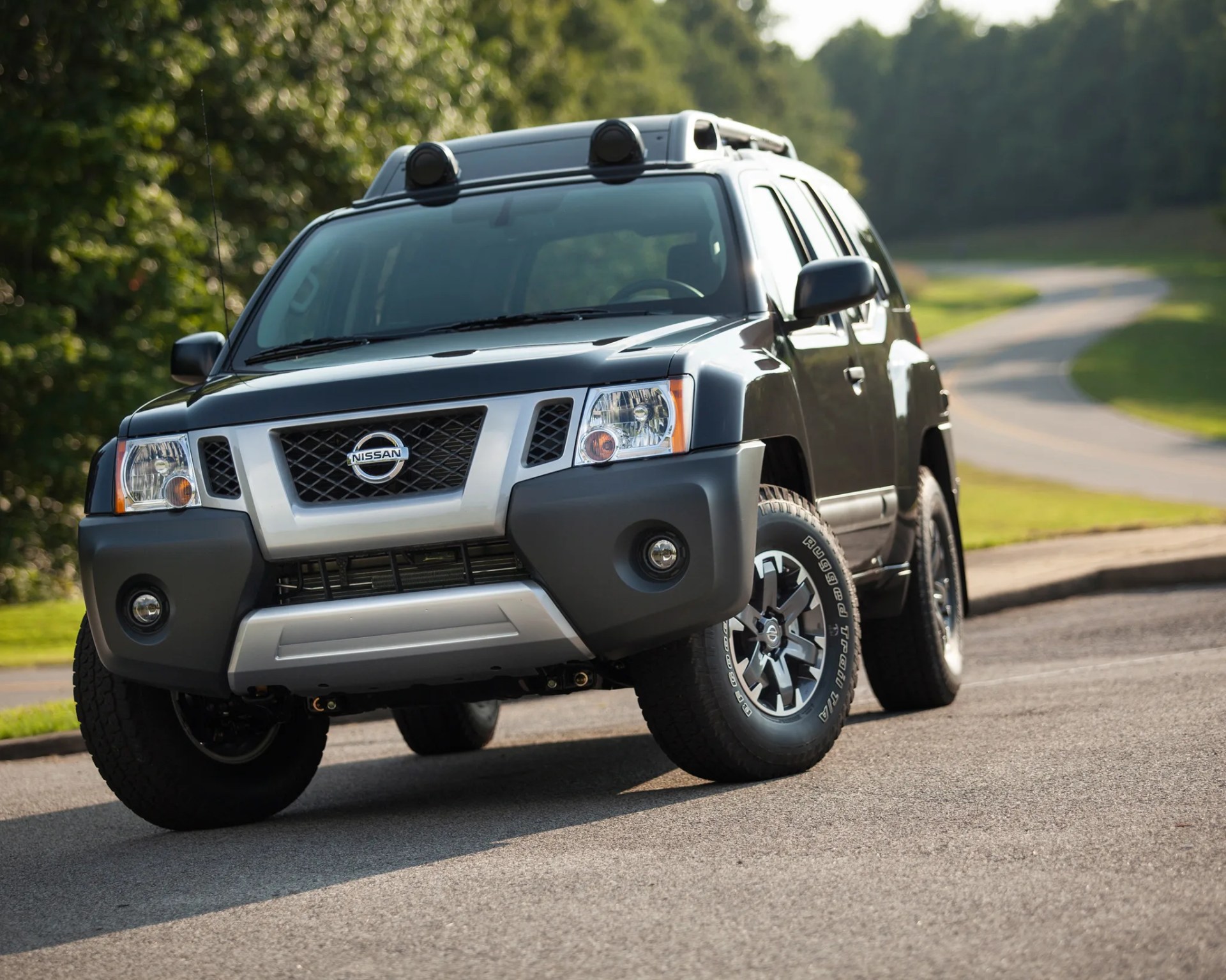 Dark gray Nissan SUV with roof lights parked on a paved road near greenery.
