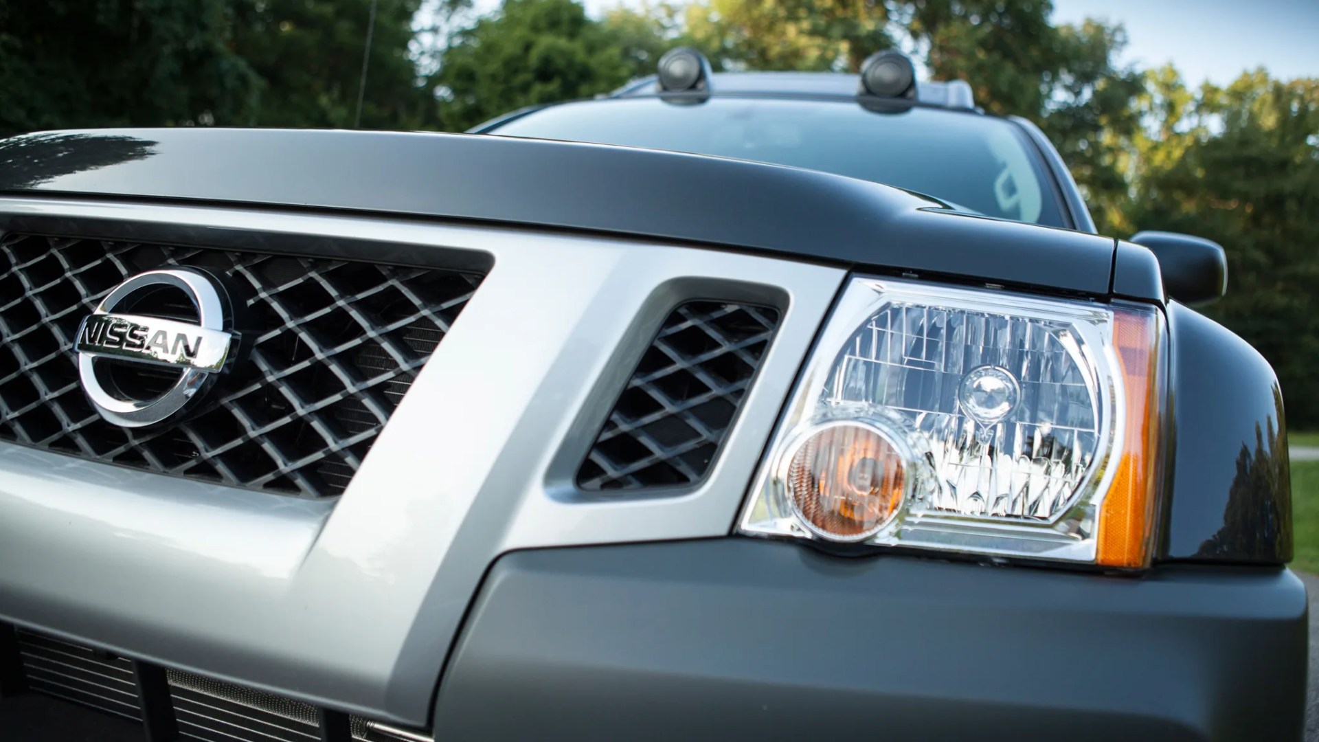 Close-up of a black and silver Nissan vehicle front grille and headlight with trees in the background.
