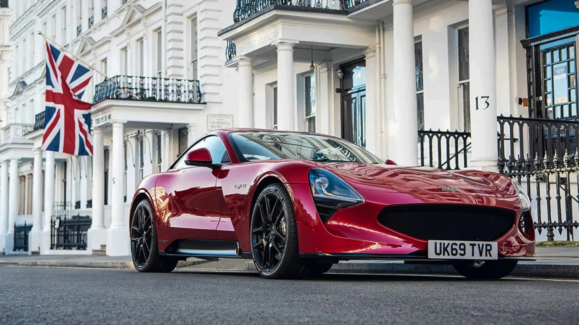Shiny red sports car with black wheels parked on a street in front of white buildings with columns and a Union Jack flag.