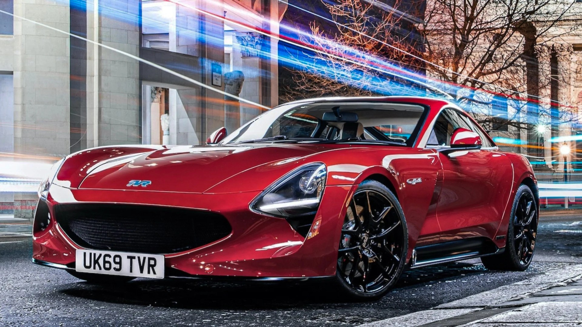 Red TVR sports car with black wheels parked on a city street at night with light trails in the background.
