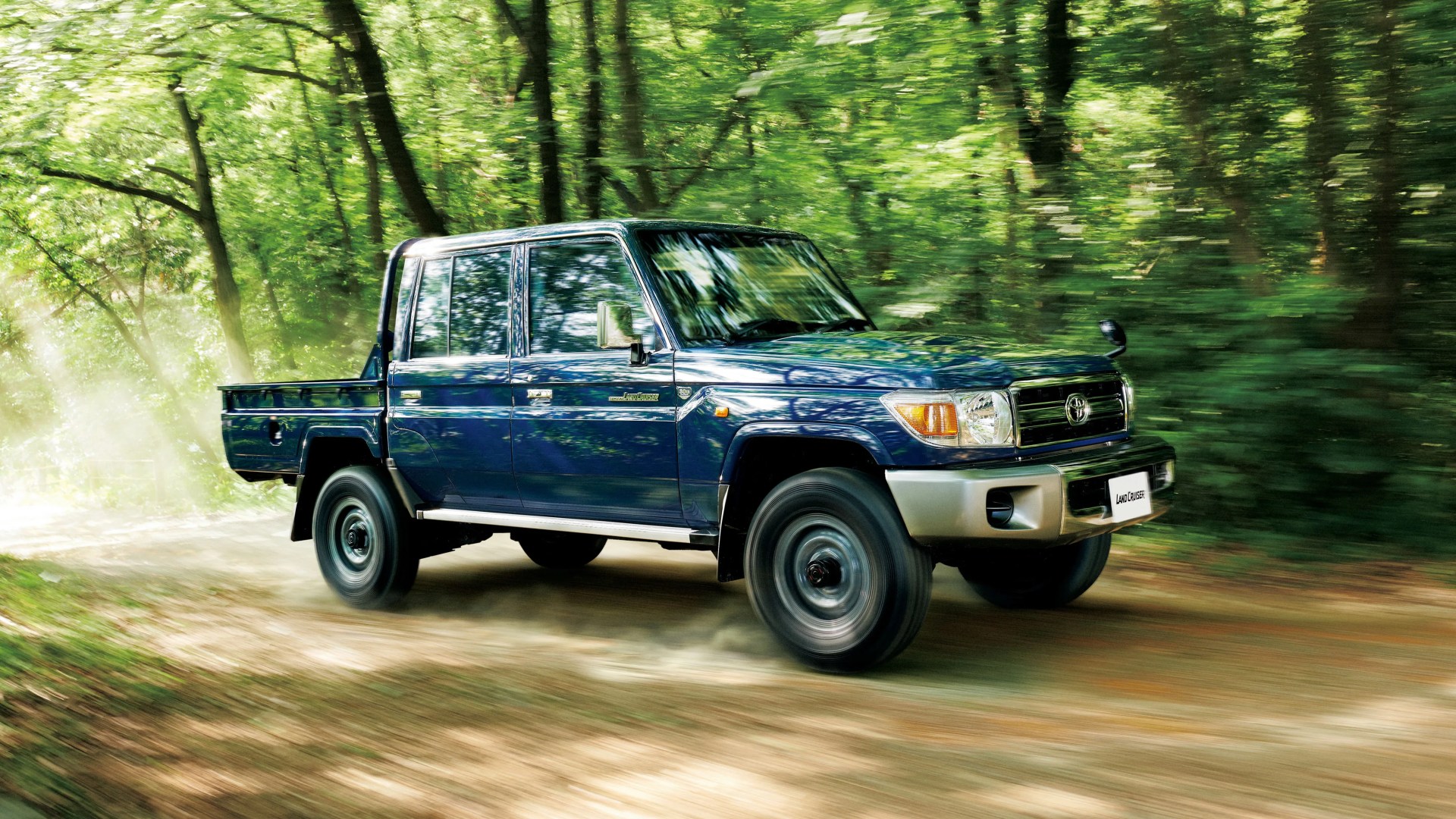 Blue Toyota Land Cruiser pickup truck driving on a dirt road through a green forest.