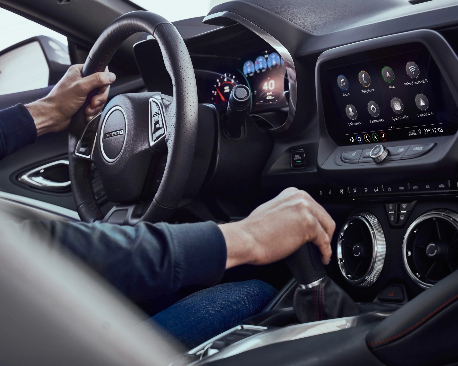 Driver's hands on black leather Camaro steering wheel and gear shift inside a car with digital dashboard and touchscreen display.