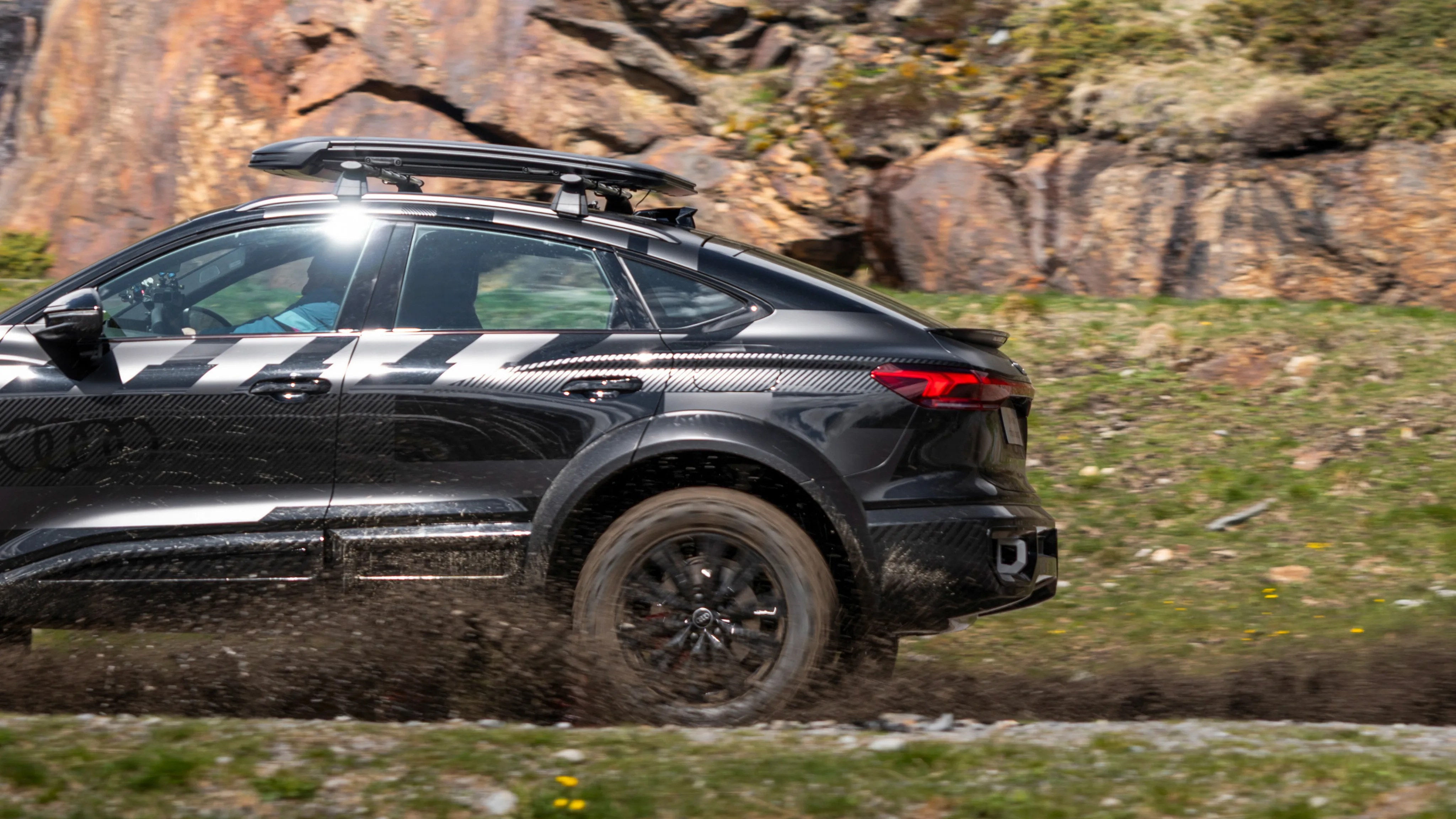 Black Audi SUV with roof rack driving on a muddy off-road trail near rocky terrain.