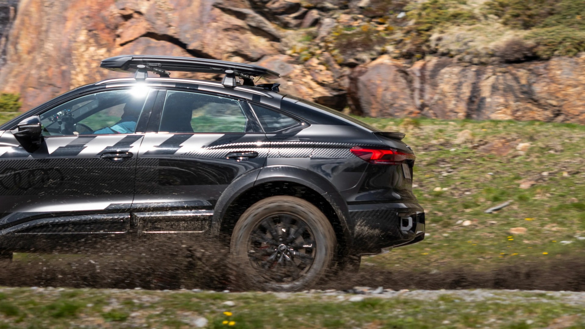 Black Audi SUV with roof rack driving on a muddy off-road trail near rocky terrain.