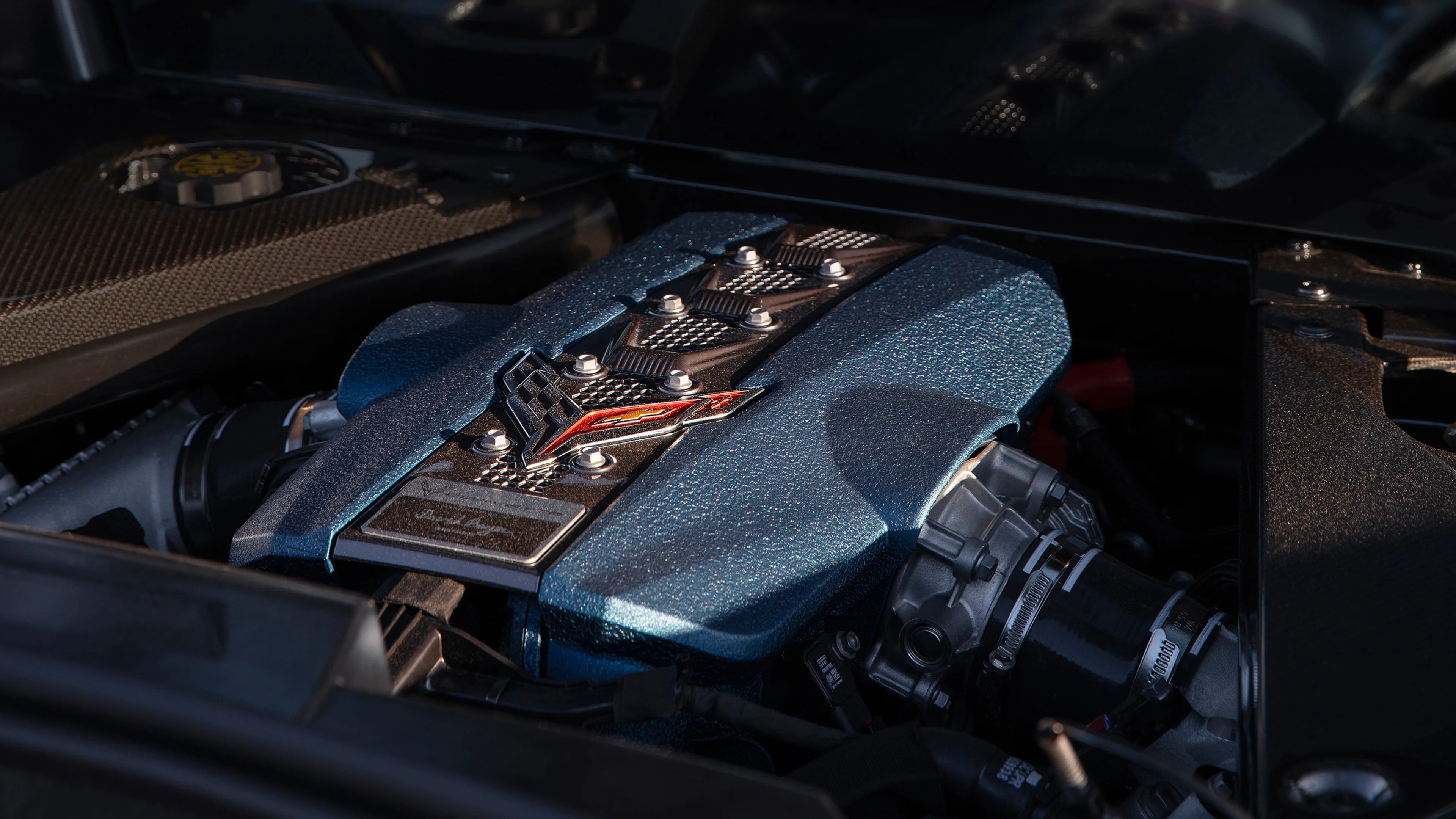 Blue textured engine cover with a red and black Corvette emblem and visible metal components around it.
