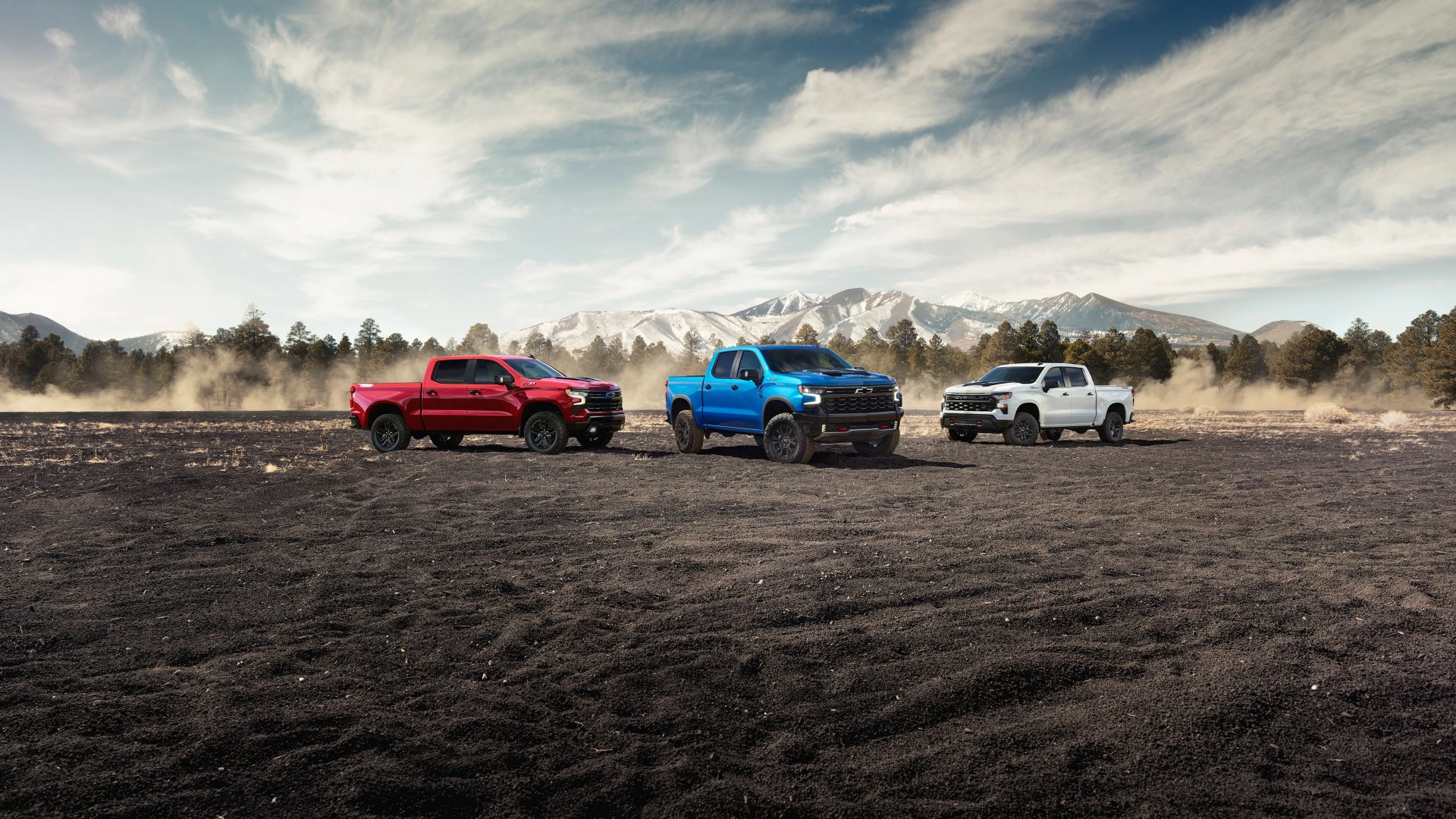 Three Chevrolet pickup trucks in red, blue, and white parked on a dirt field with mountains and trees in the background.
