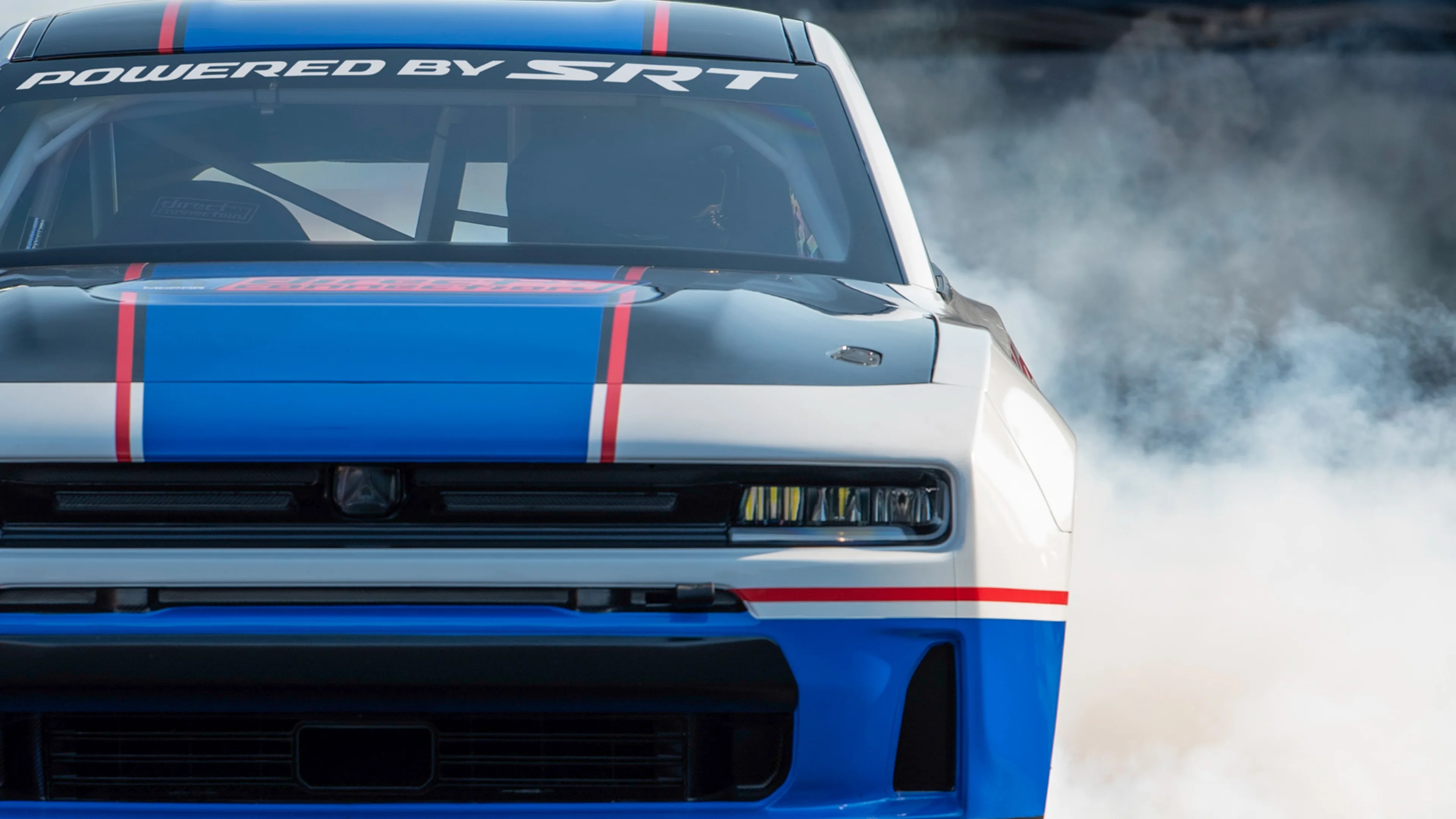 Front view of a white, blue, and black race car with "Powered by SRT" decal on the windshield and smoke in the background.