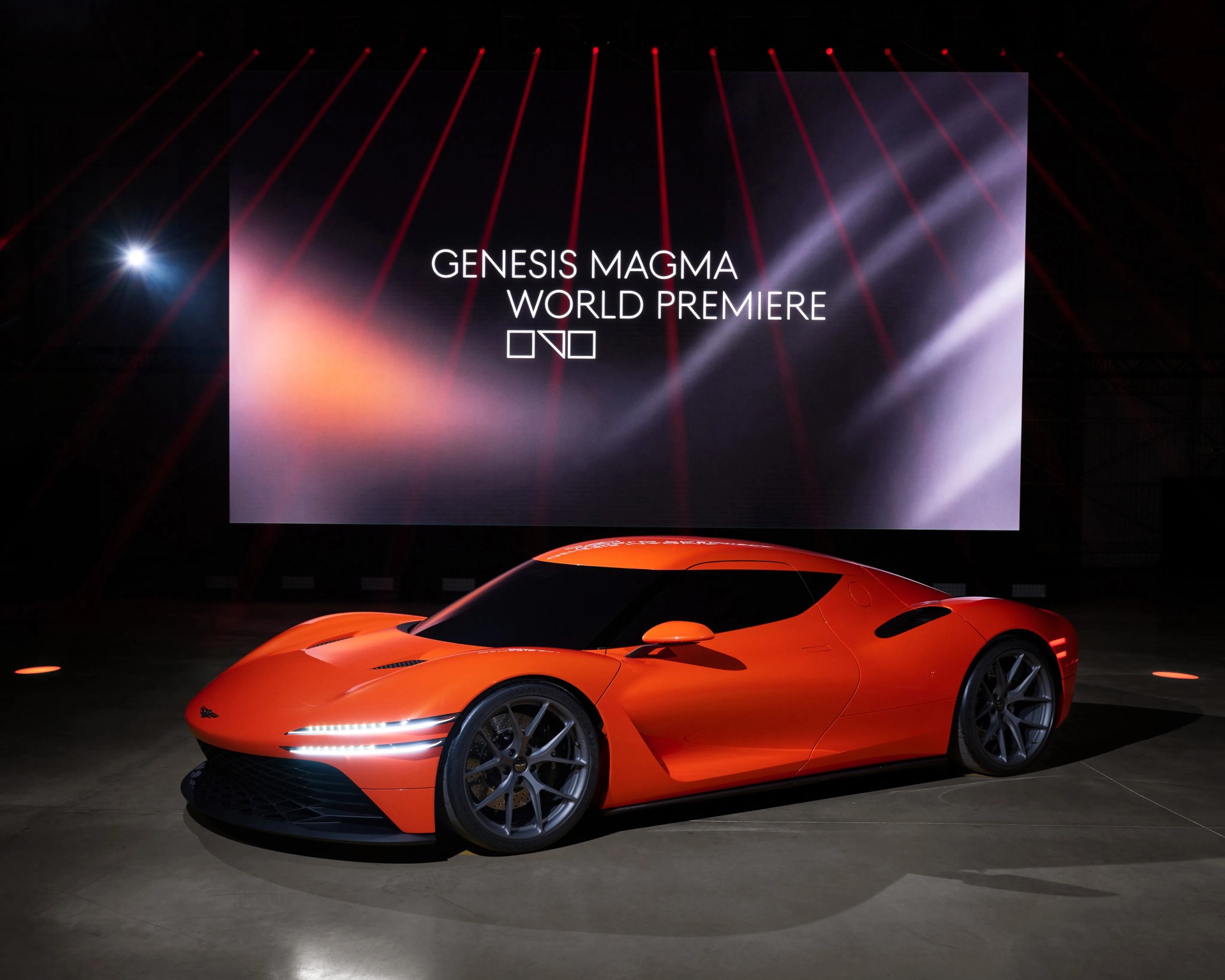 Bright orange Genesis Magma sports car with sleek design and LED headlights in a dark showroom with a large screen behind.