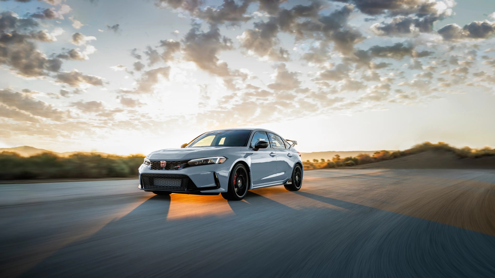 Gray Honda Civic Type R hatchback driving on a road at sunset with a cloudy sky.