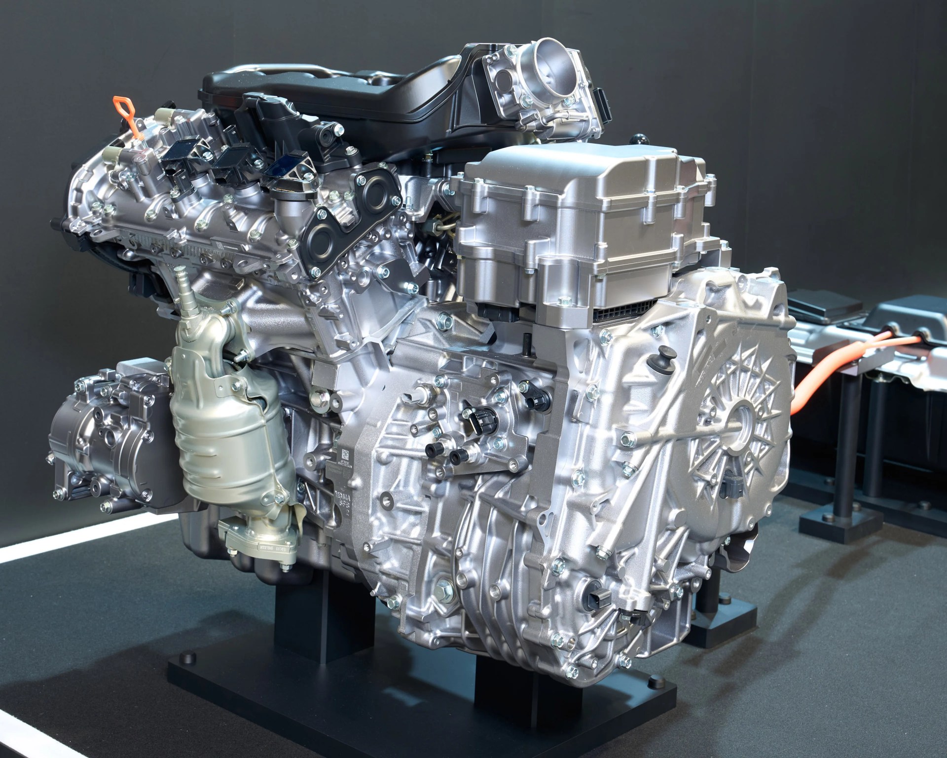Silver and black hybrid car engine with orange high-voltage cable on a black display stand.