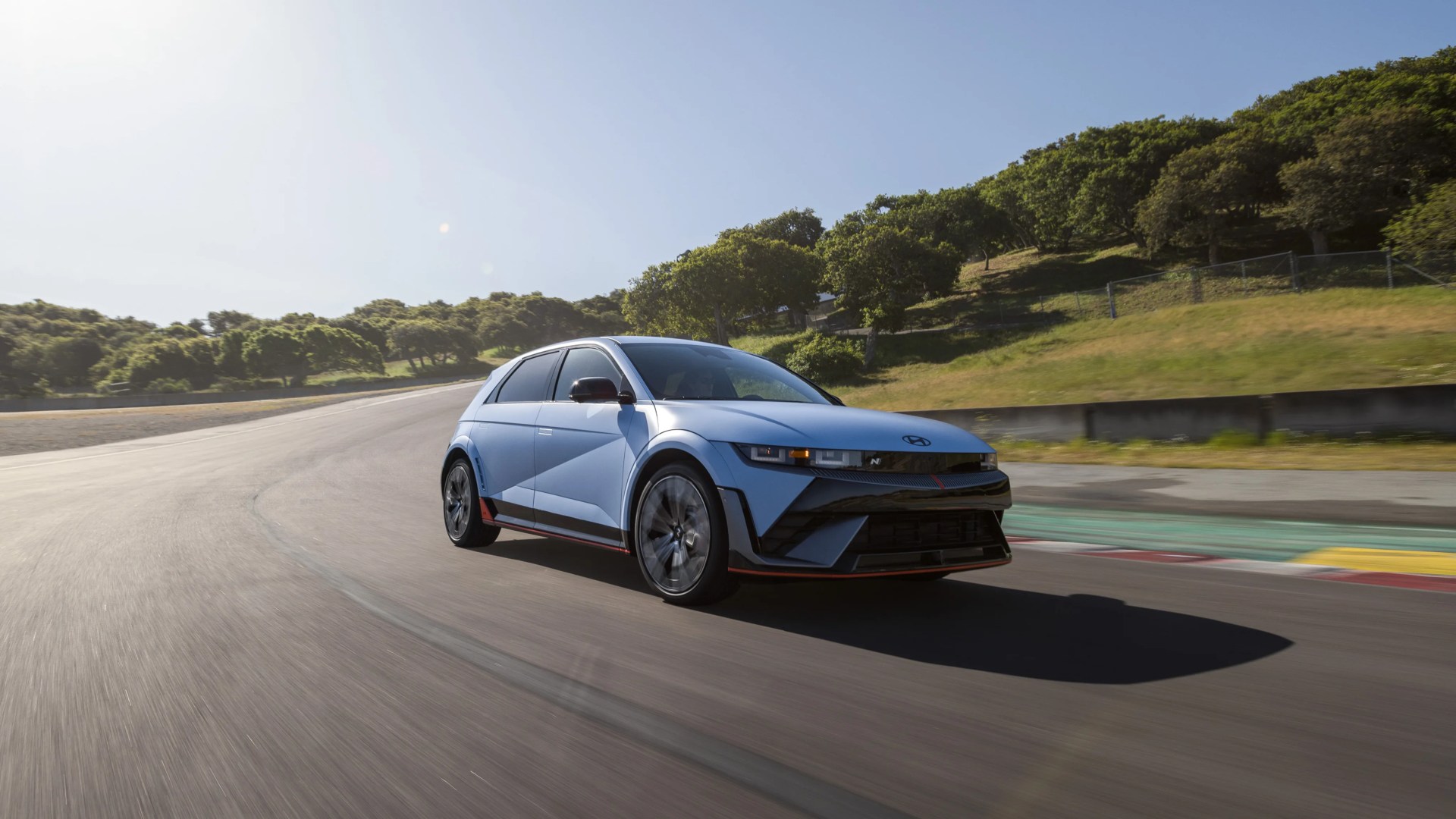 Light blue Hyundai electric hatchback driving on a sunny racetrack with green trees in the background.