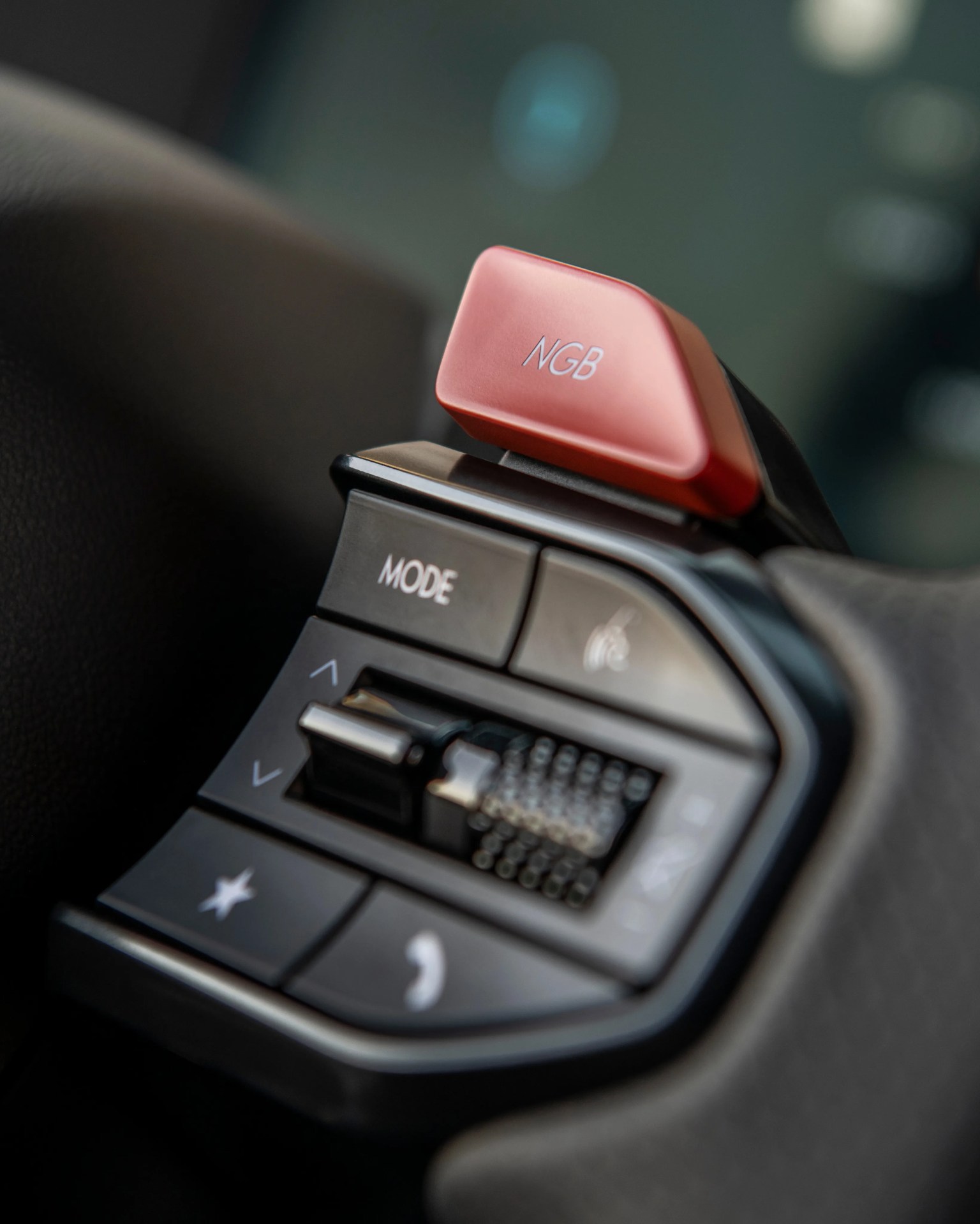 Close-up of a car steering wheel control panel with buttons labeled "MODE," volume up/down arrows, a star, a phone icon, and a red paddle shifter marked "NGB.