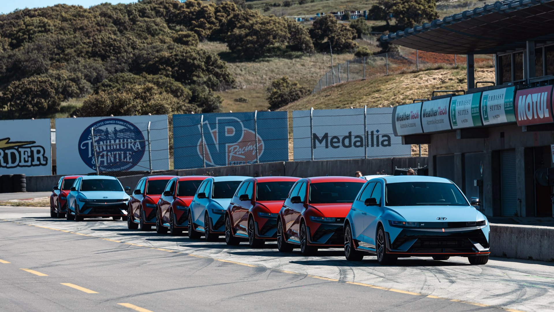 Seven Hyundai Ioniq 5 electric cars in alternating red and light blue colors lined up on a racetrack pit lane.