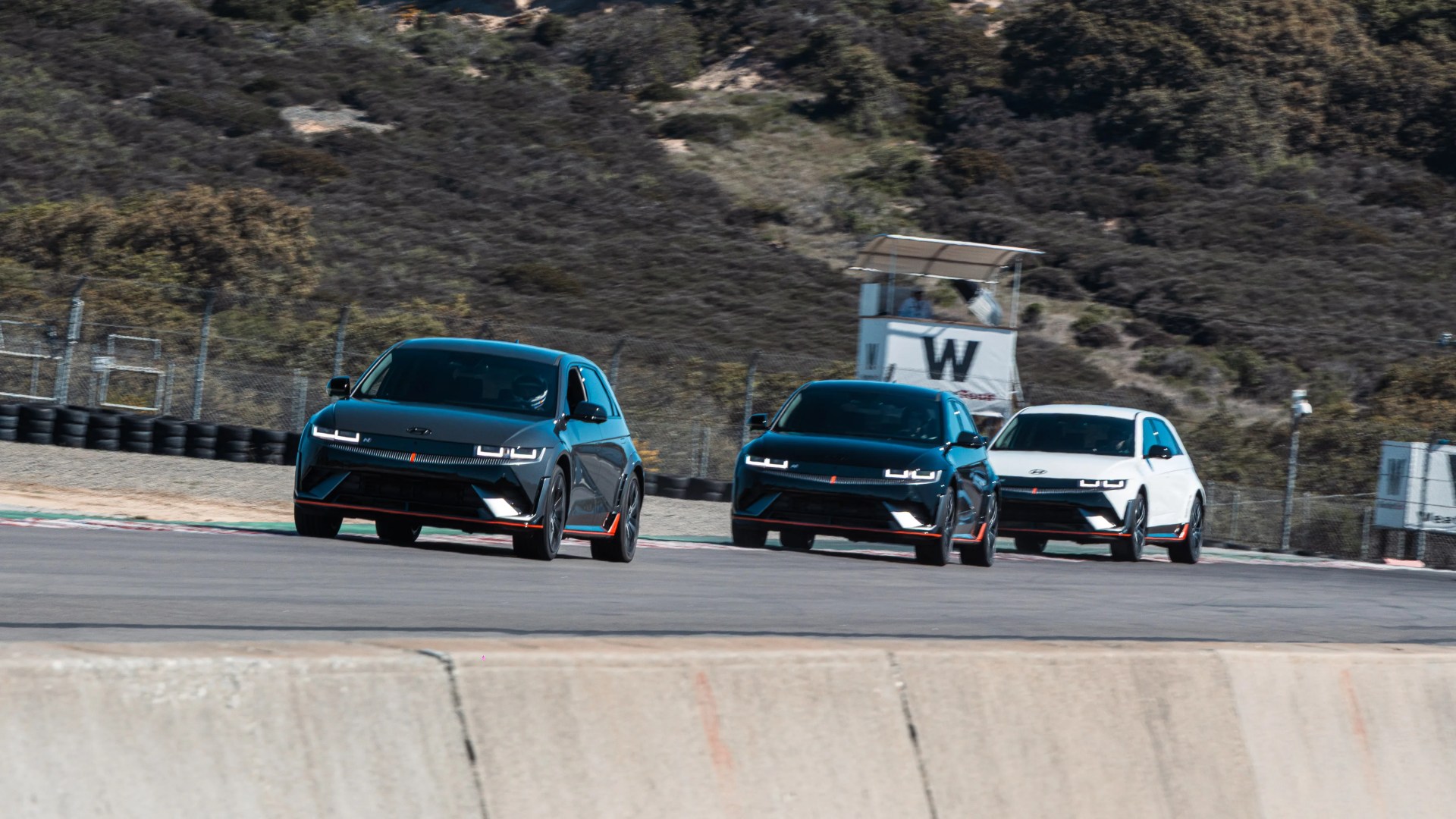 Three Hyundai electric hatchbacks, two black and one white, driving on a race track with hills in the background.