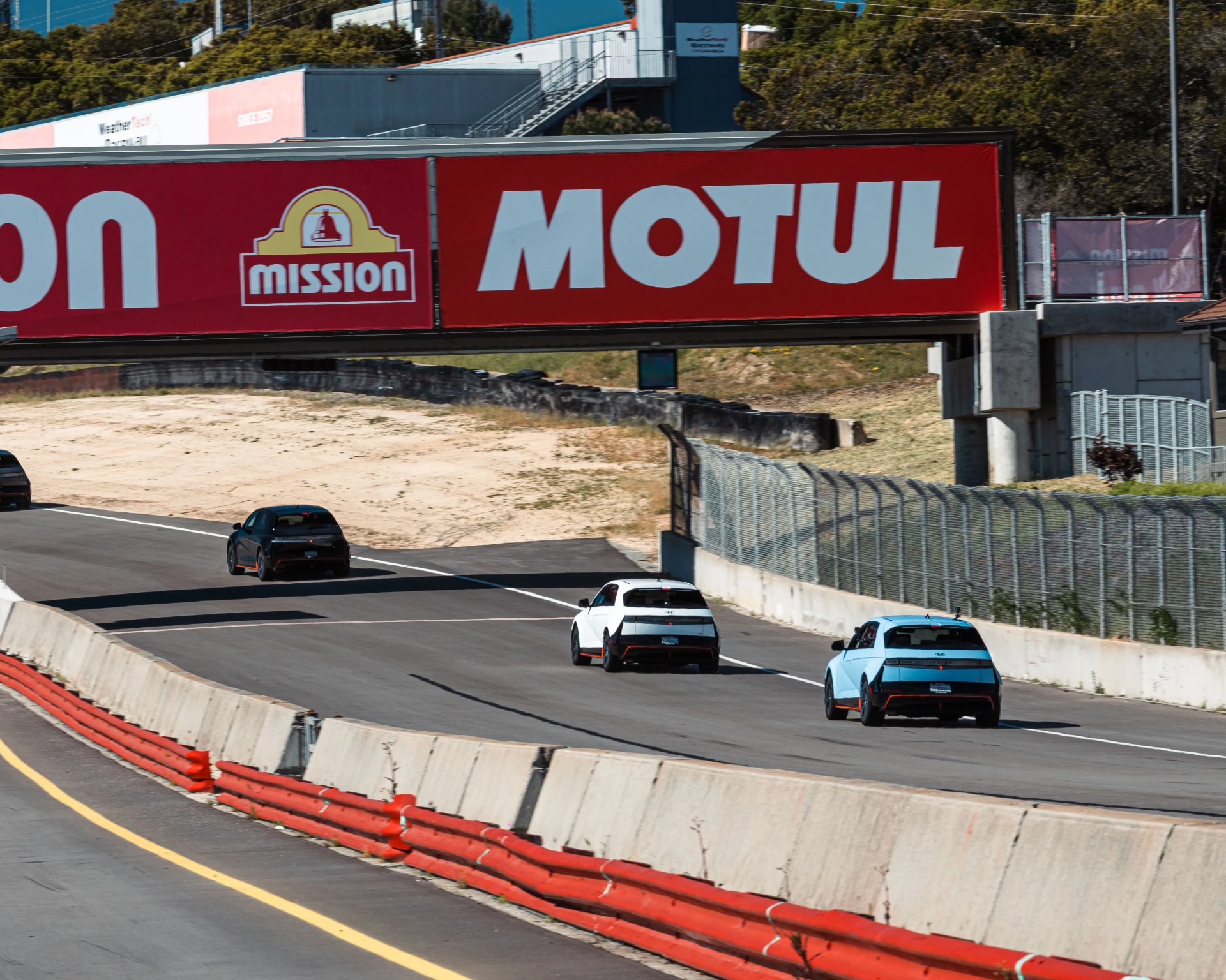 Three compact electric cars, two white and one light blue, driving on a racetrack under a red "MOTUL" banner.