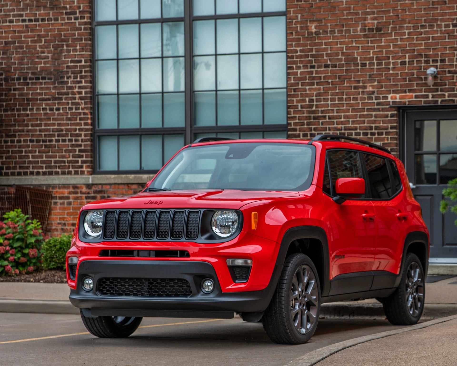 Red Jeep Renegade SUV parked on a street in front of a brick building with large windows.