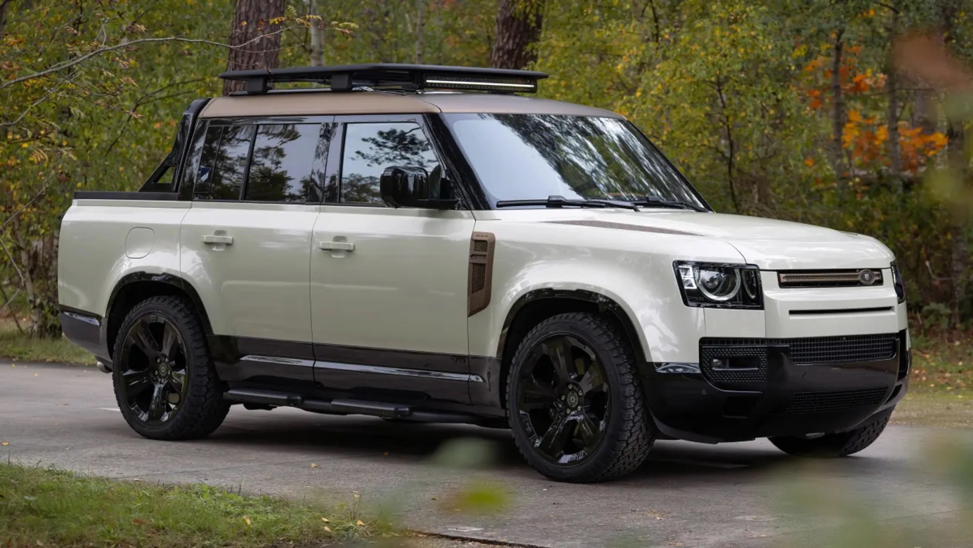 White and black Land Rover Defender pickup truck with roof rack parked on a forest road.