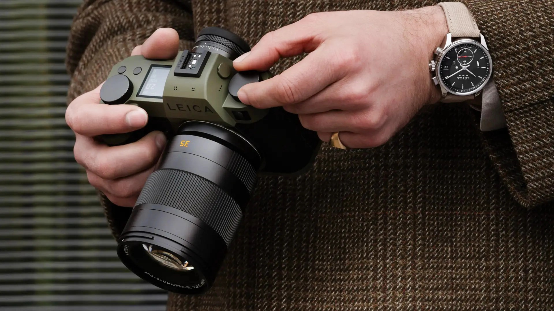 Person holding an olive-green Leica camera with a black 35mm lens, wearing a black Leica wristwatch with a beige strap.