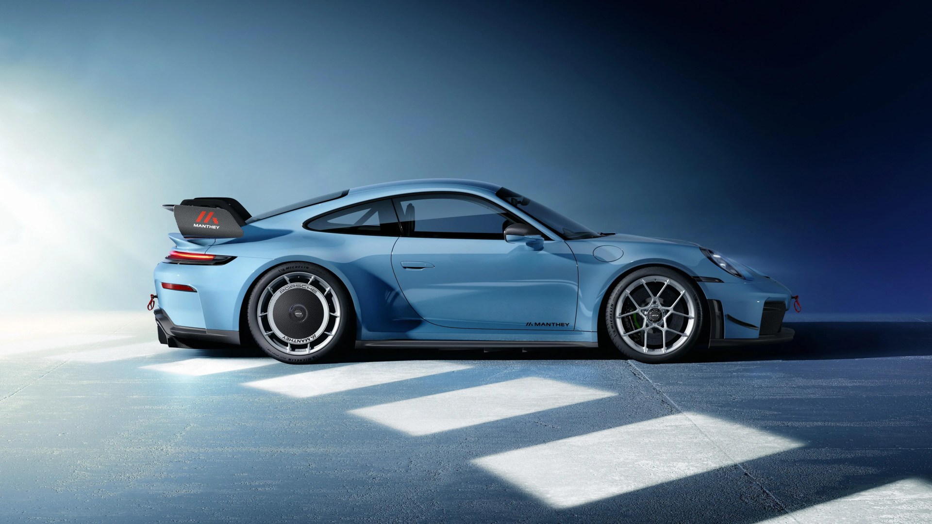 Light blue Manthey Porsche sports car with aerodynamic rear wing and unique wheel covers on a blue and white floor.