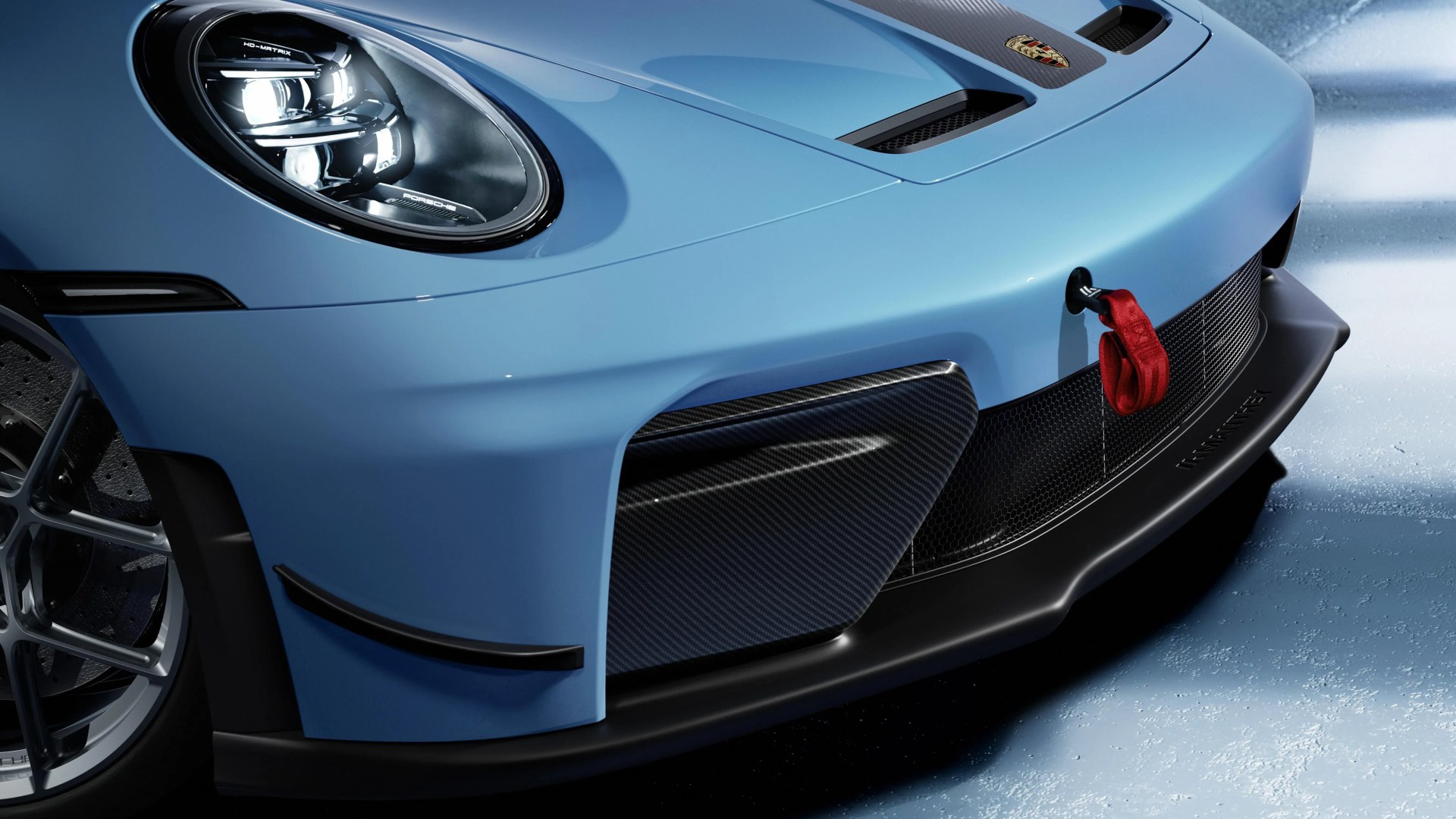 Front corner of a light blue Porsche sports car showing headlight, carbon fiber vent, red tow strap, and black front splitter.