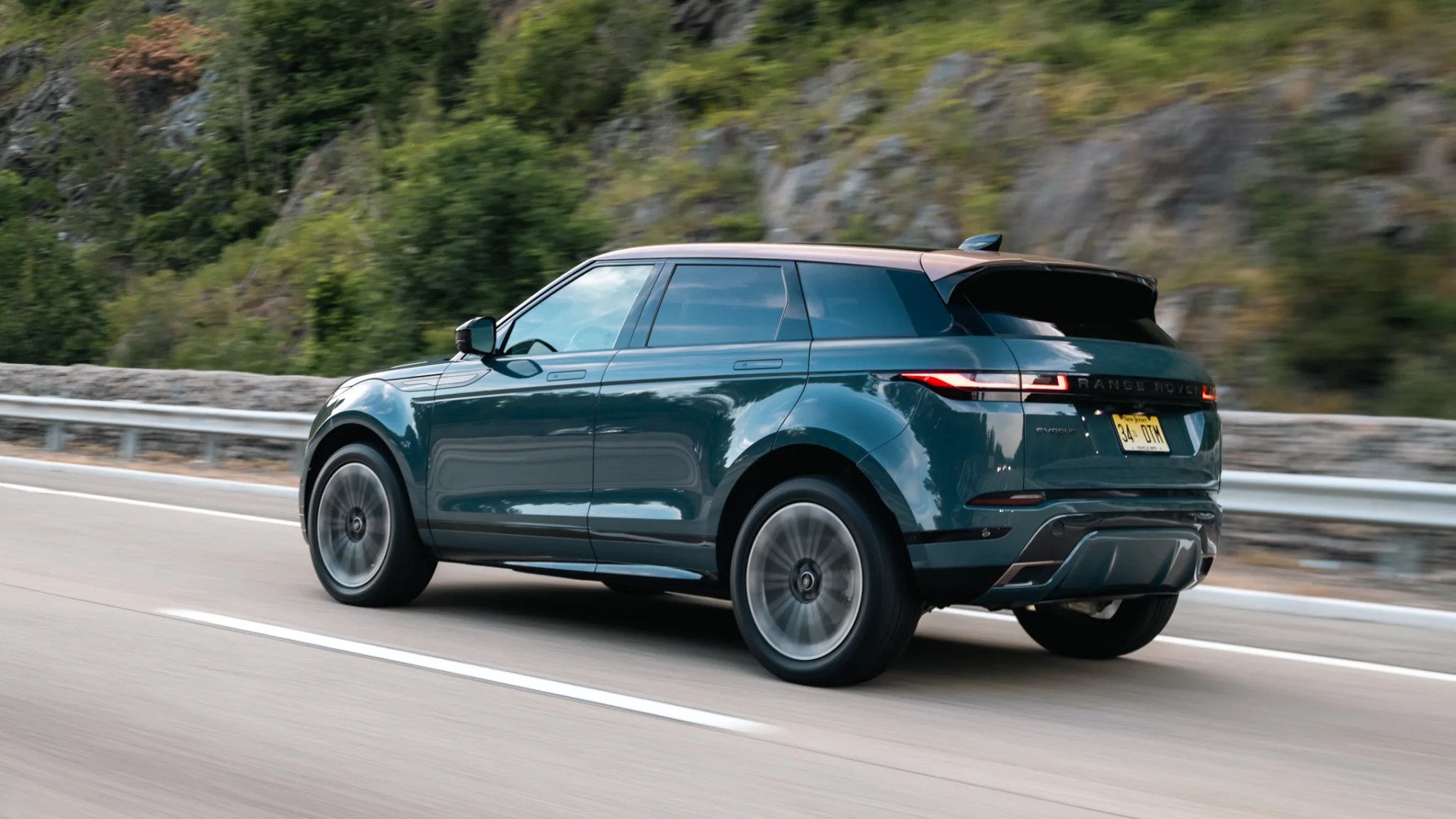 Dark green Range Rover Evoque SUV driving on a highway with blurred wheels and forest background.