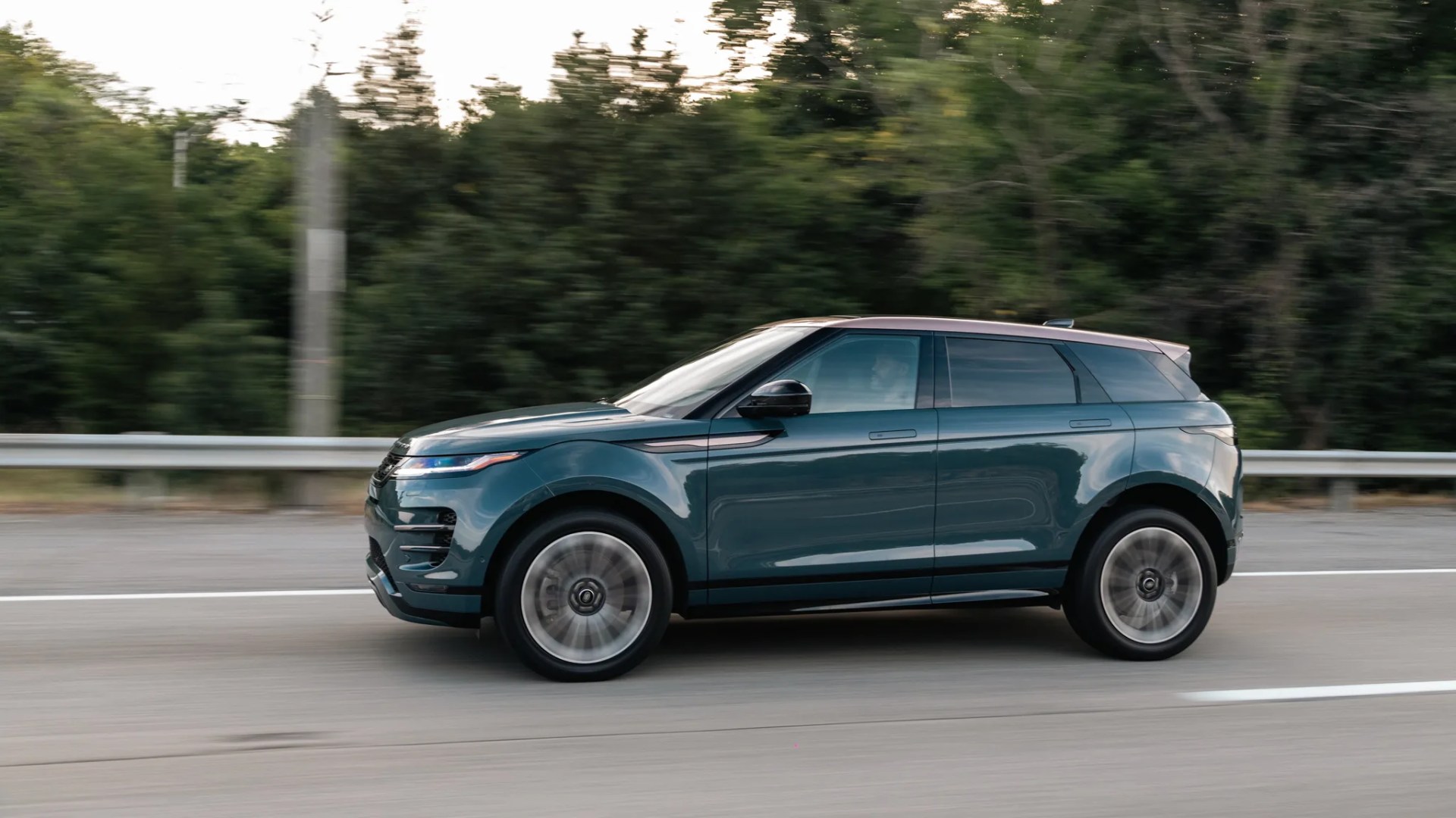 Dark teal compact SUV with black roof driving on a road with blurred background trees.