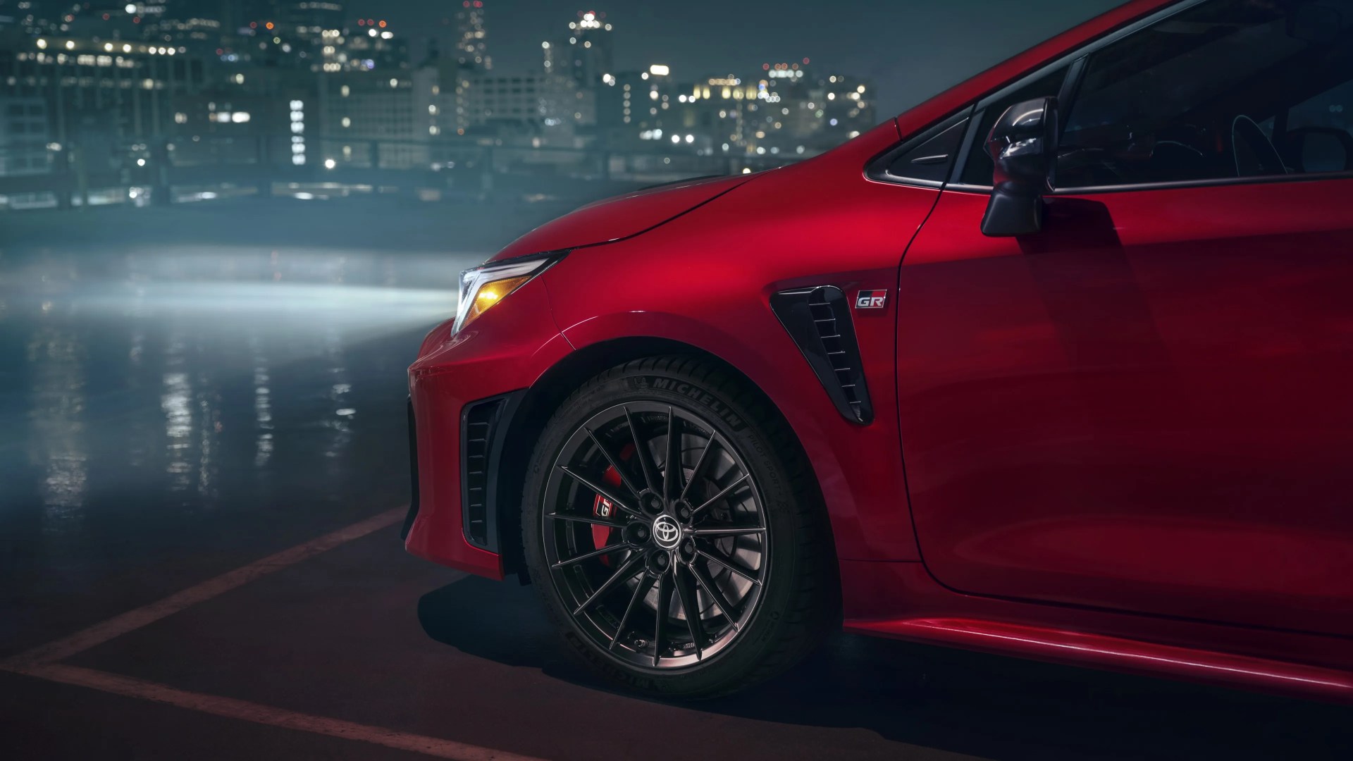 Front side of a red Toyota GR car with black alloy wheel and illuminated headlight at night.