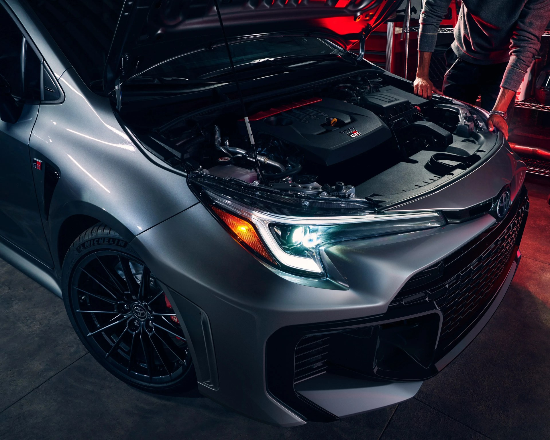 Silver Toyota GR car with open hood showing engine and black alloy wheels in a garage.