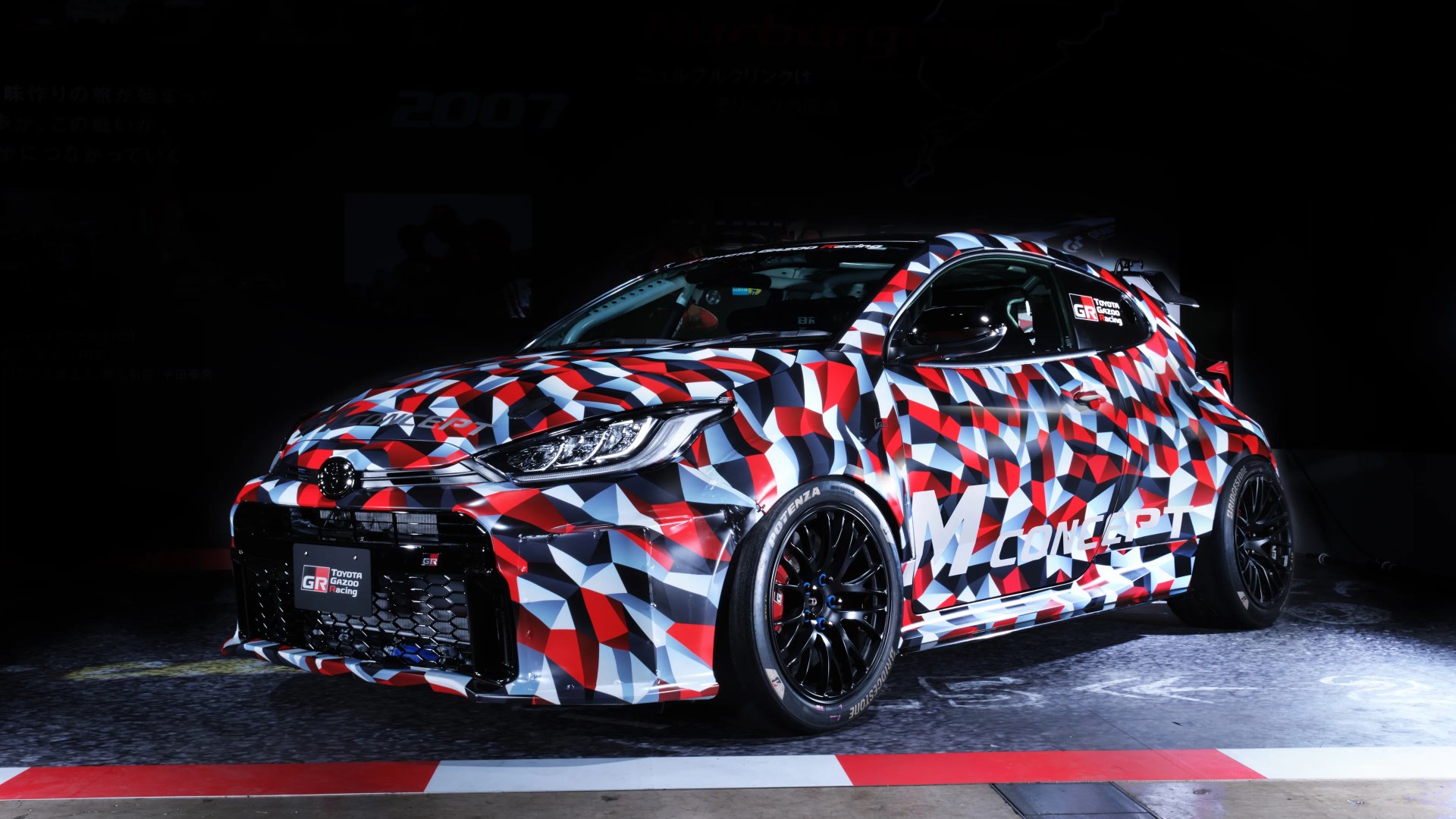 Toyota GR racing car with red, white, and black geometric camouflage and black wheels on a dark floor.