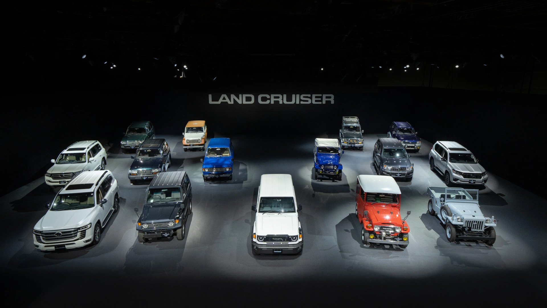 Multiple Toyota Land Cruiser models in various colors displayed in a dark room with "LAND CRUISER" text on the back wall.