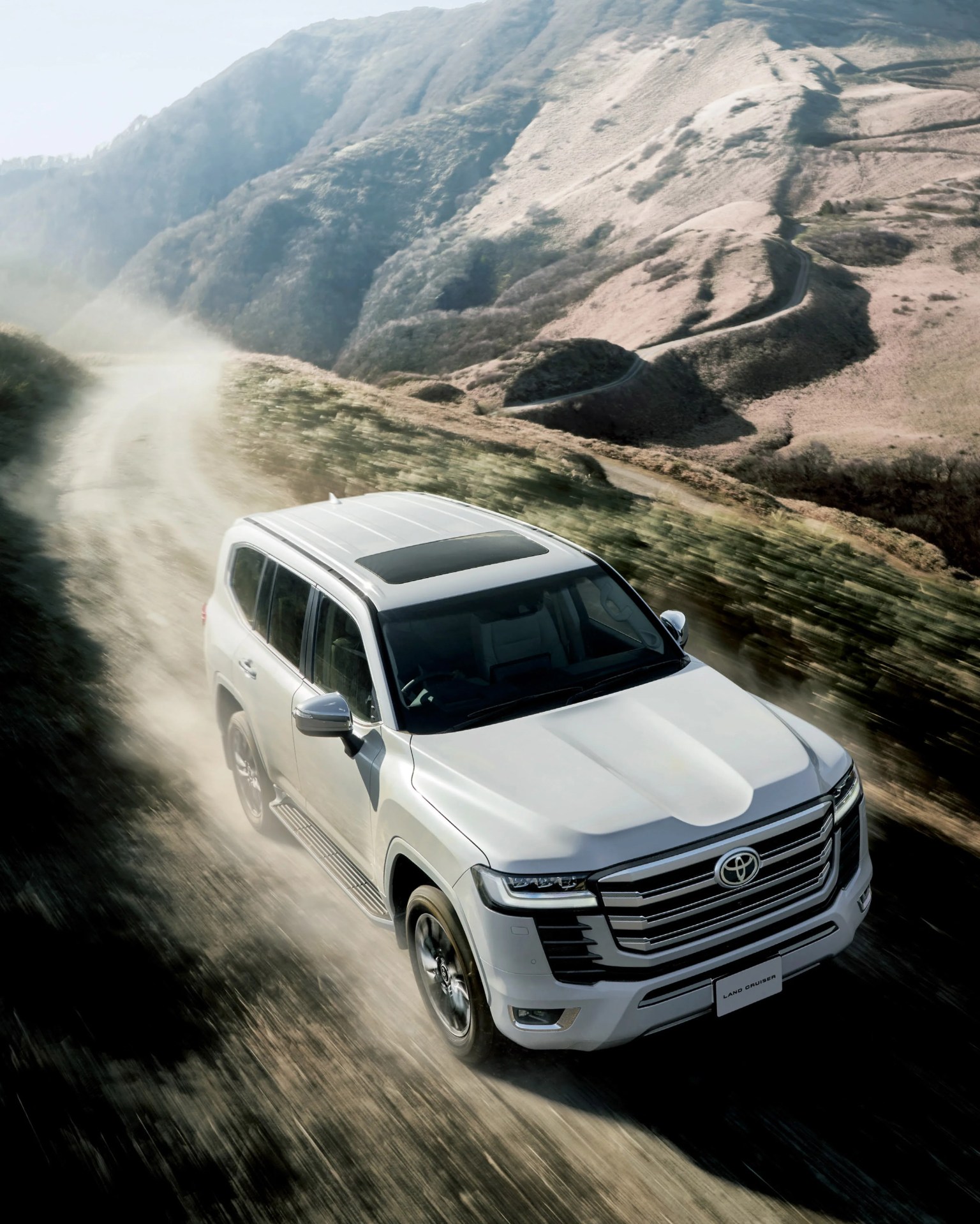 White Toyota Land Cruiser SUV driving on a dusty mountain dirt road with rugged hills in the background.