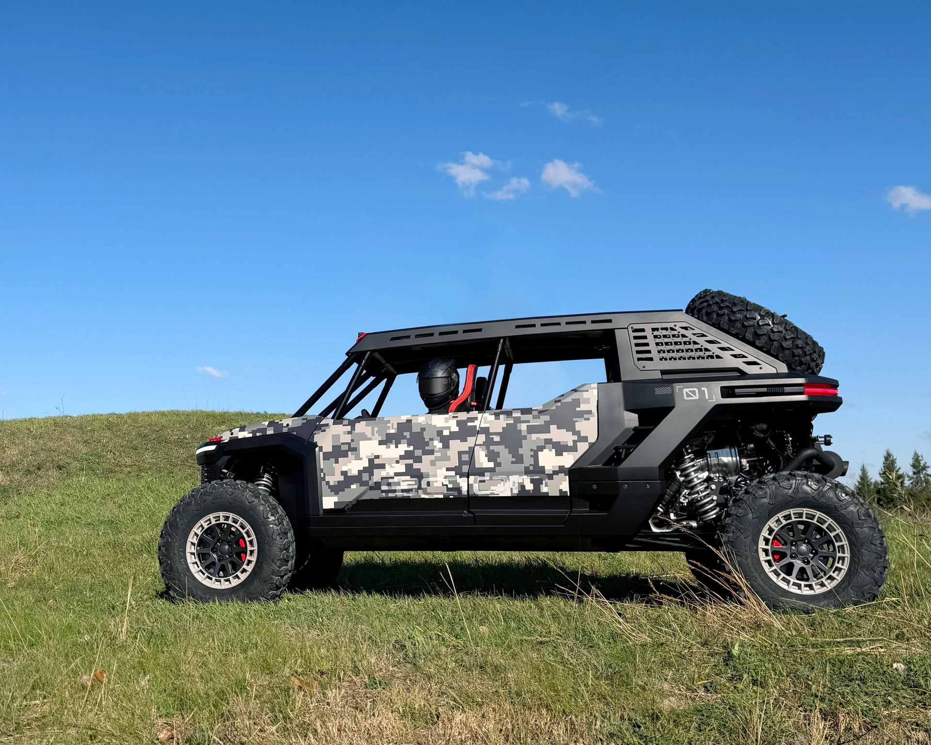 Off-road vehicle with digital camouflage body and large rugged tires parked on grass under clear blue sky.